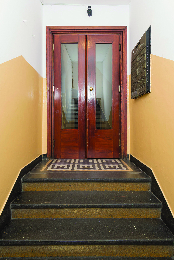 Strefa wejściowa w czynszowym domu wielorodzinnym przy ul. Słupeckiej 9, posadzka wykonana z lastryka, przed drzwiami kompozycja z gorsecik&oacute;w, na ścianie historyczna skrzynka na listy.