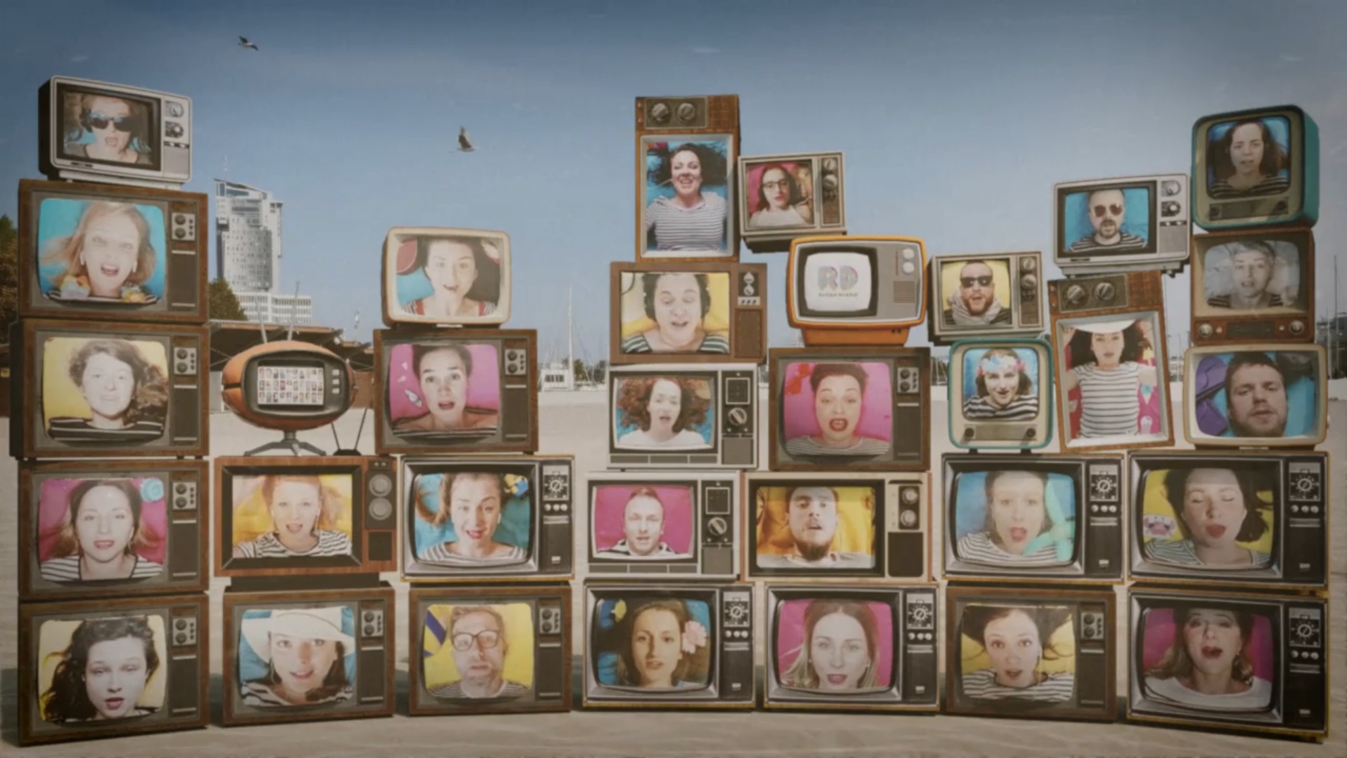 Kadr z teledysku ch&oacute;ru Razzle Dazzle do utworu The Beach Boys - "Barbar' Ann". Poszczeg&oacute;lni ch&oacute;rzyści zaprezentowani w ekranach telewizor&oacute;w, w tle gdyńska plaża i budynek Sea Towers oraz niebo i latające mewy // materiały prasowe