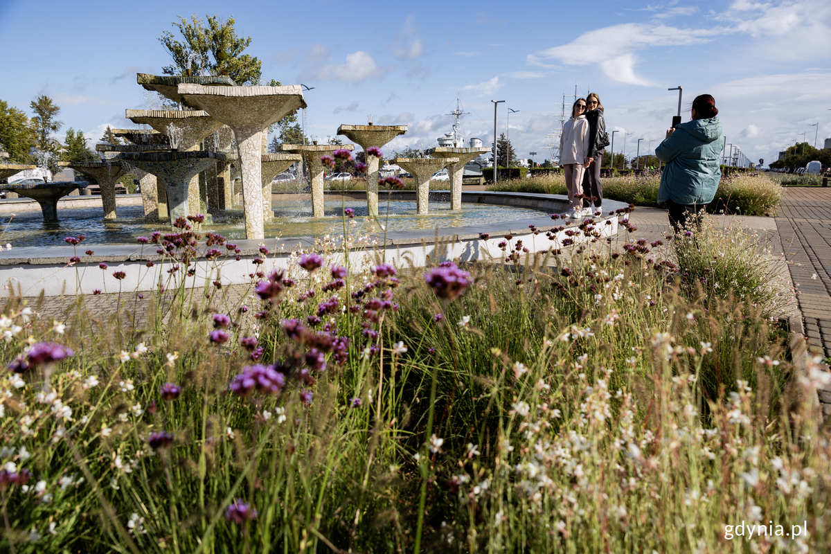 Dwie kobiety pozuja do zdjęcia przy fontannie, wok&oacute;ł rosna rośliny.