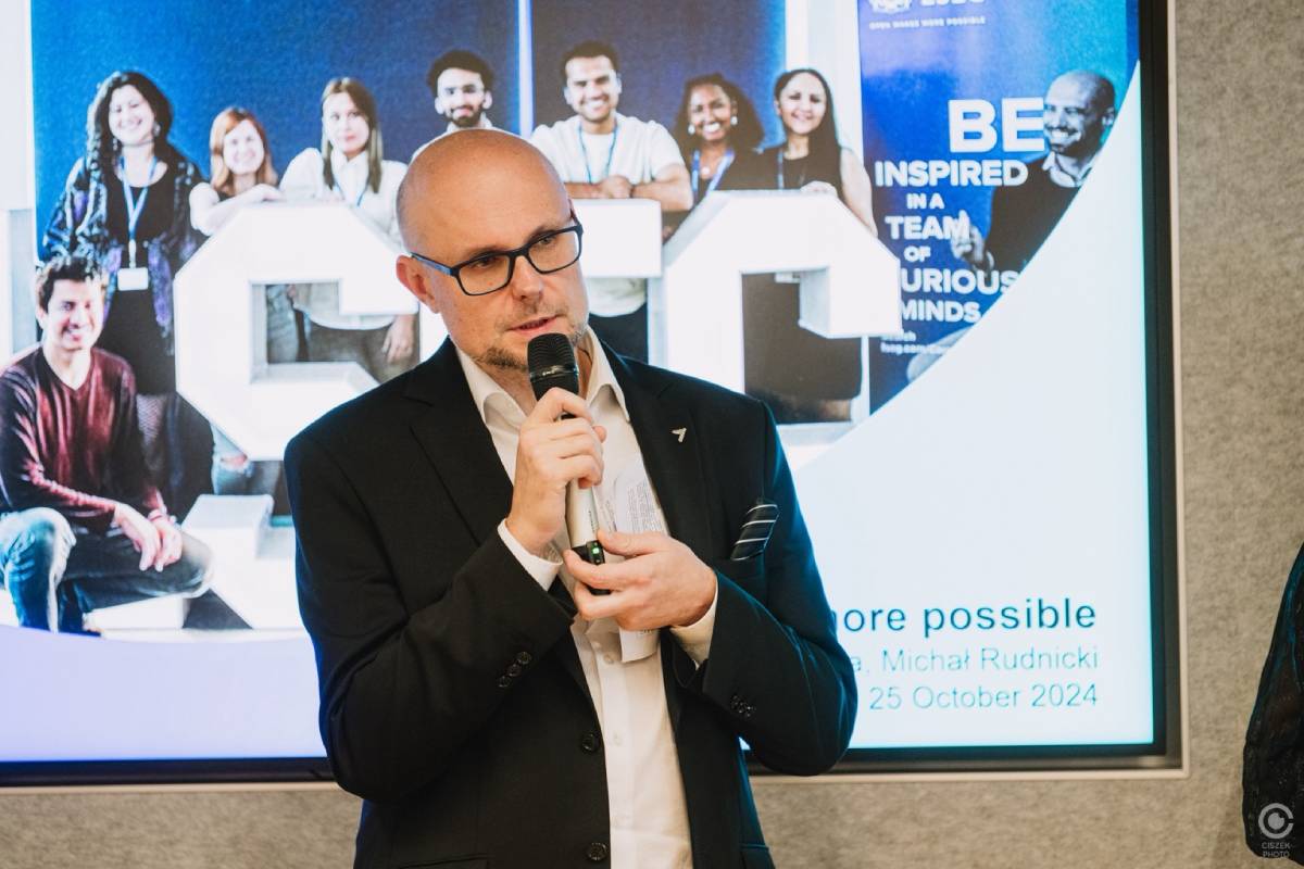 Elegancko ubrany mężczyzna m&oacute;wiący do mikrofonu na tle grafiki przedstawiającej grupę os&oacute;b