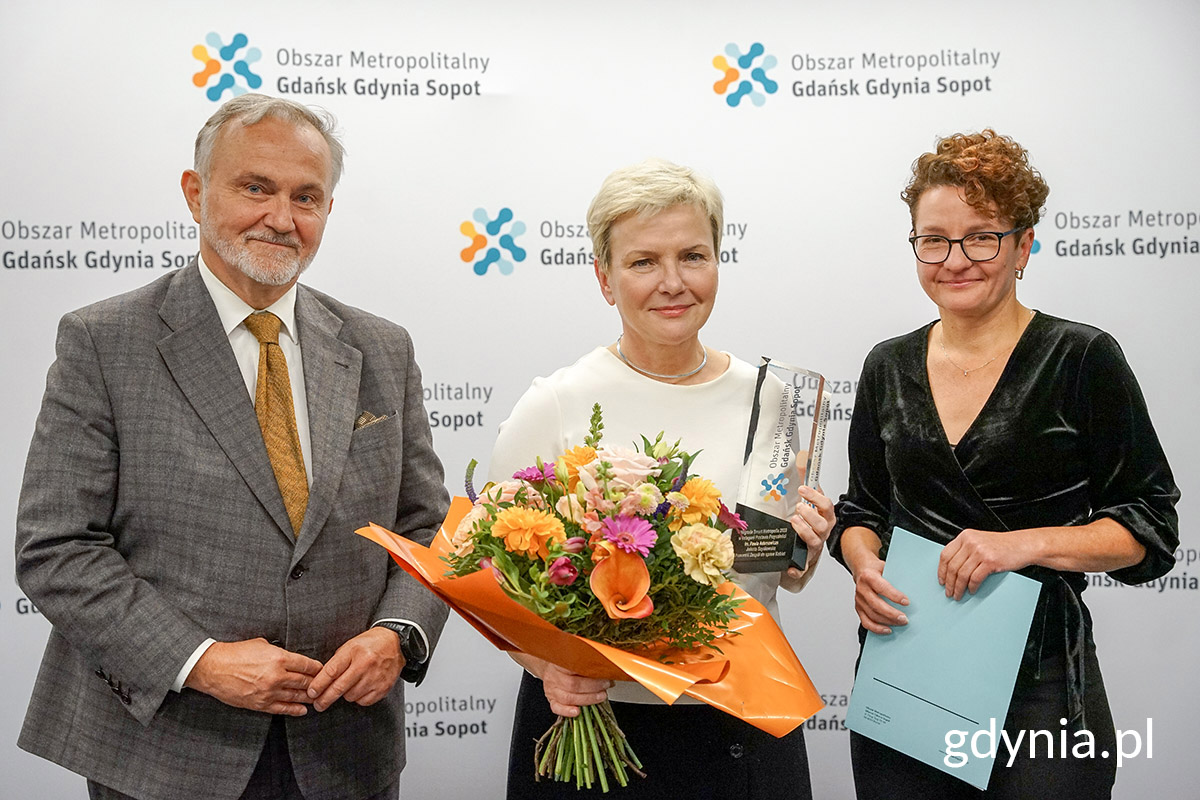 Wojciech Szczurek, Jolanta Szydłowska i Magdalena Czarzyńska-Jachim pozują do zdjęcia. W tle biała ścianka OMGGS, kobieta w środku trzyma nagrodę i kwiaty
