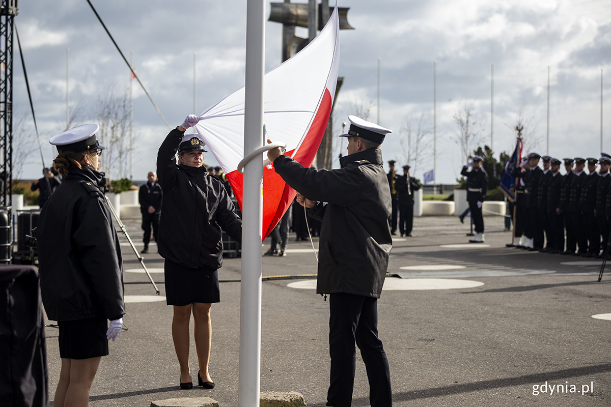 Kobieta i mężczyzna w mundurach marynarskich wciągają flagę Polski na maszt.