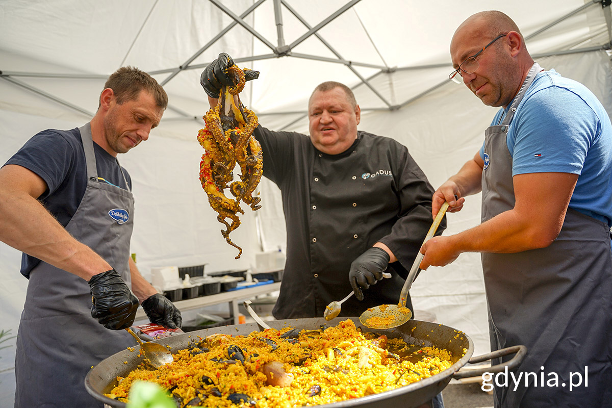 Kucharz w towarzystwie dw&oacute;ch innych trzyma nad patelnią z potrawą ośmiornicę