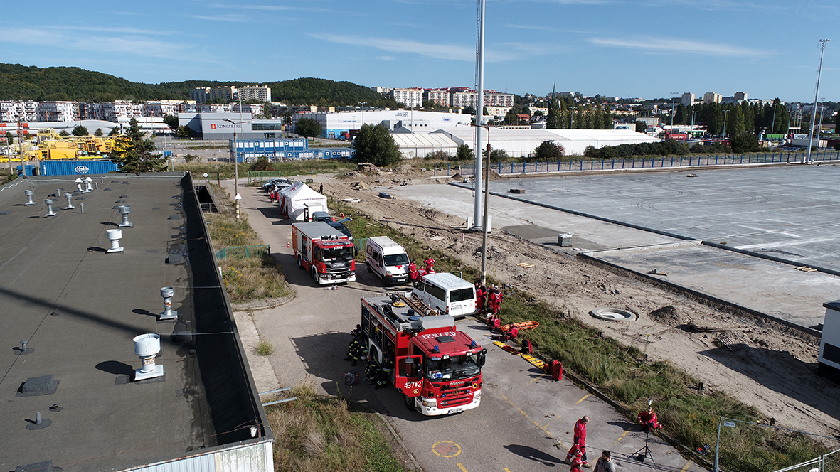 Manewry ratownicze w Porcie Gdynia. Zdjęcie robione z g&oacute;ry, z drona, przy budynku zaparkowane są wozy strażackie.