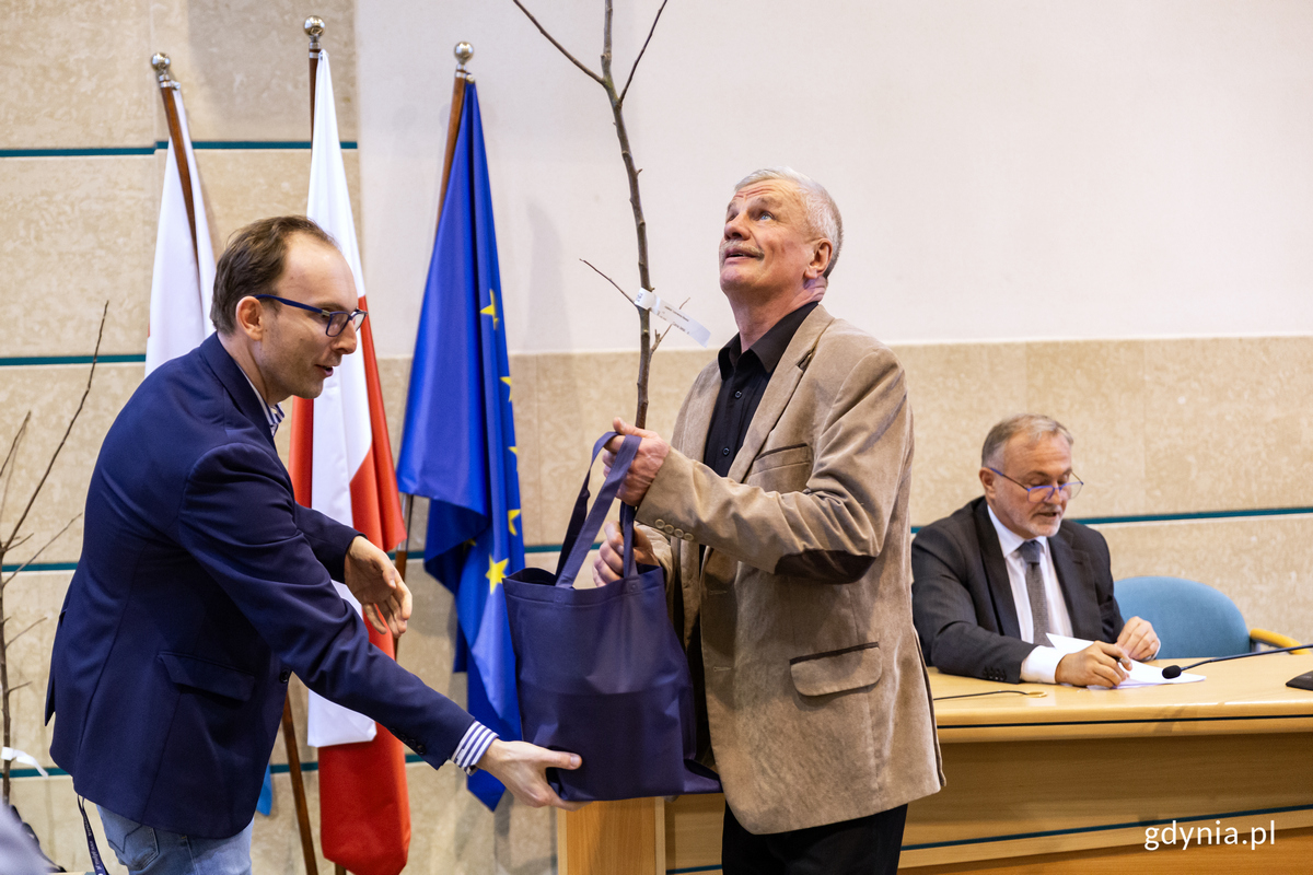 Mężczyzna wręcza mężczyźnie torbę z sadzonkja jabłoni, w tle ludzie podpisują umowy na realizacje projekt&oacute;w działkowych.