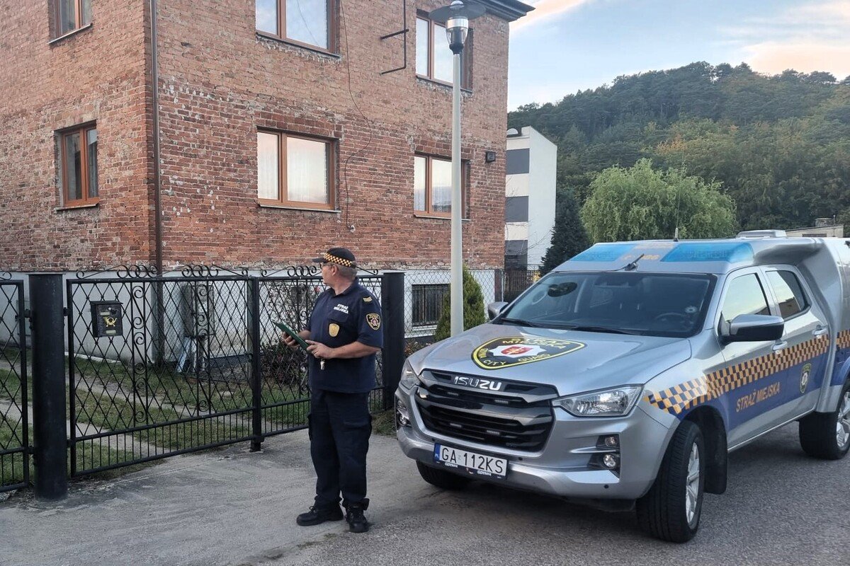 Ulica, dom, radiow&oacute;z straży miejskiej w Gdyni, obok stoi strażnik w mundurze, patrzy w kierunku domu, trzyma dokumenty