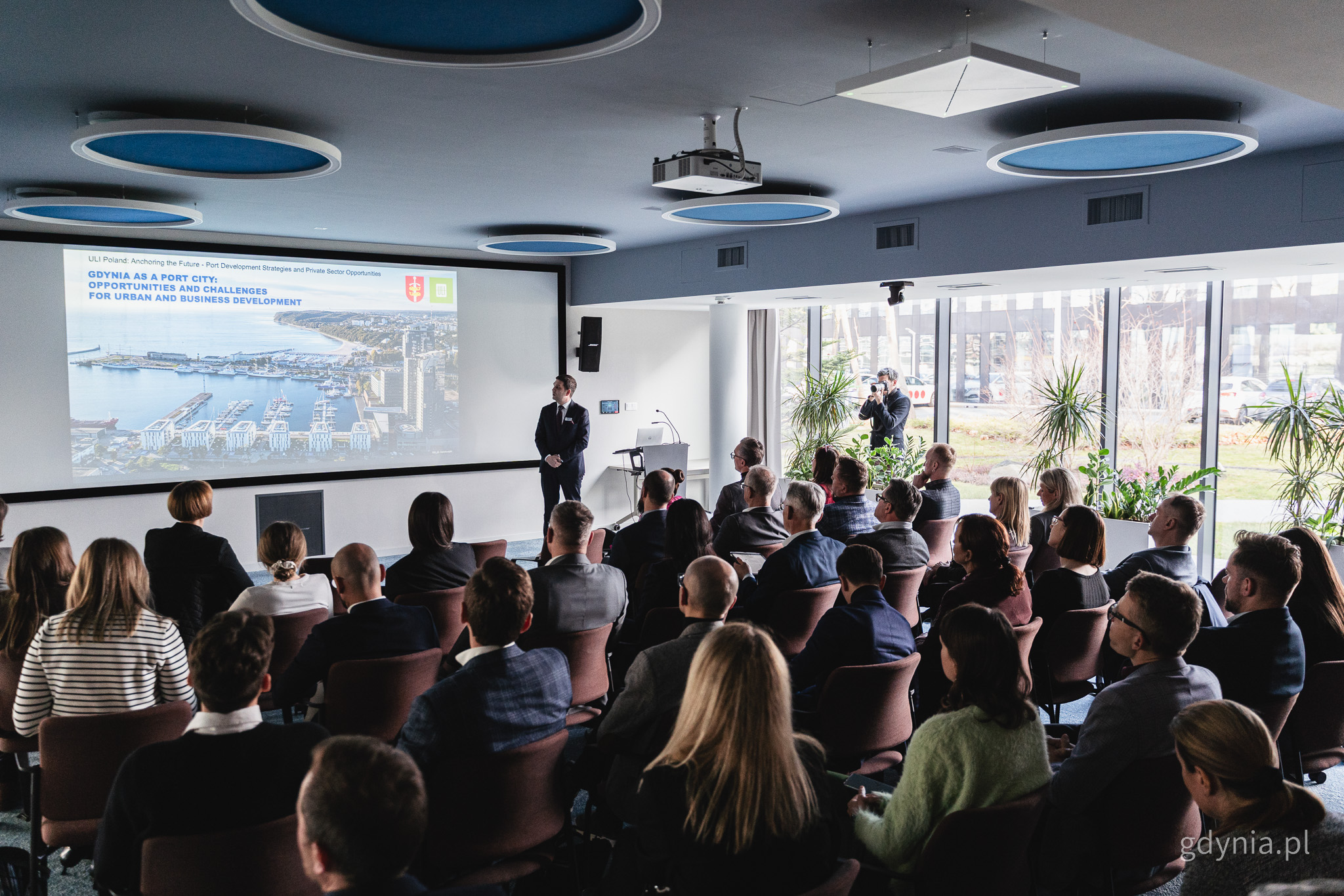 Sala konferencyjna, ludzie na widowni, przemawia wiceprezydent Maciej Zielonka, wyświetlany ekran