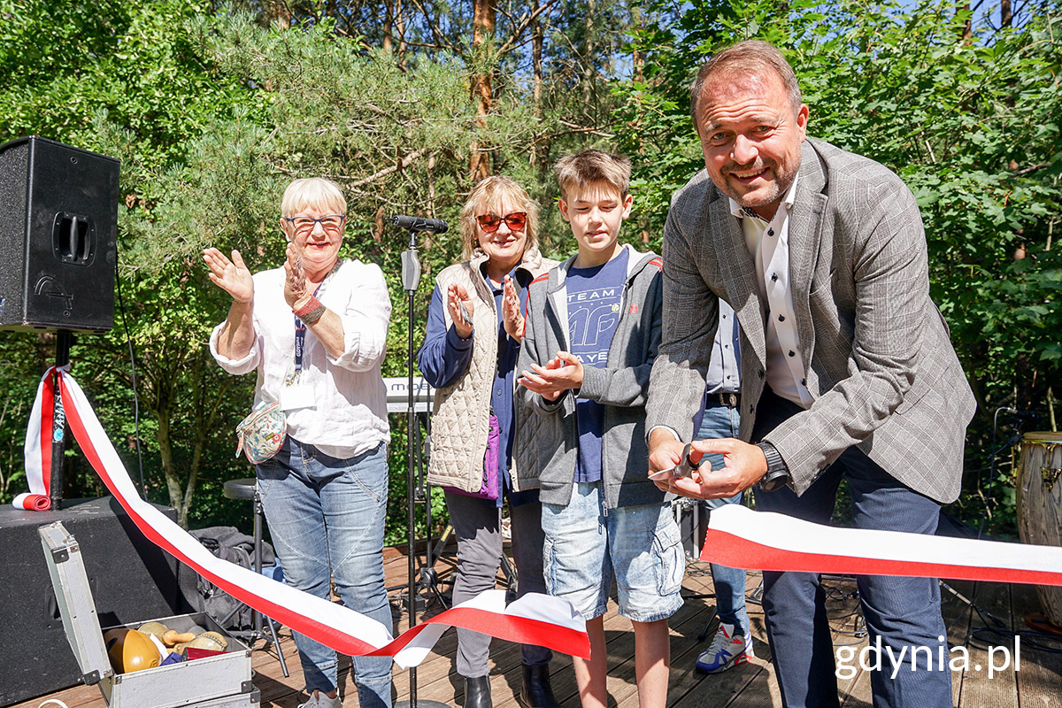 Kilka os&oacute;b w rzędzie, biało-czerwona wstęga, Marek Łucyk przecina wstęgę, za nimi widać park