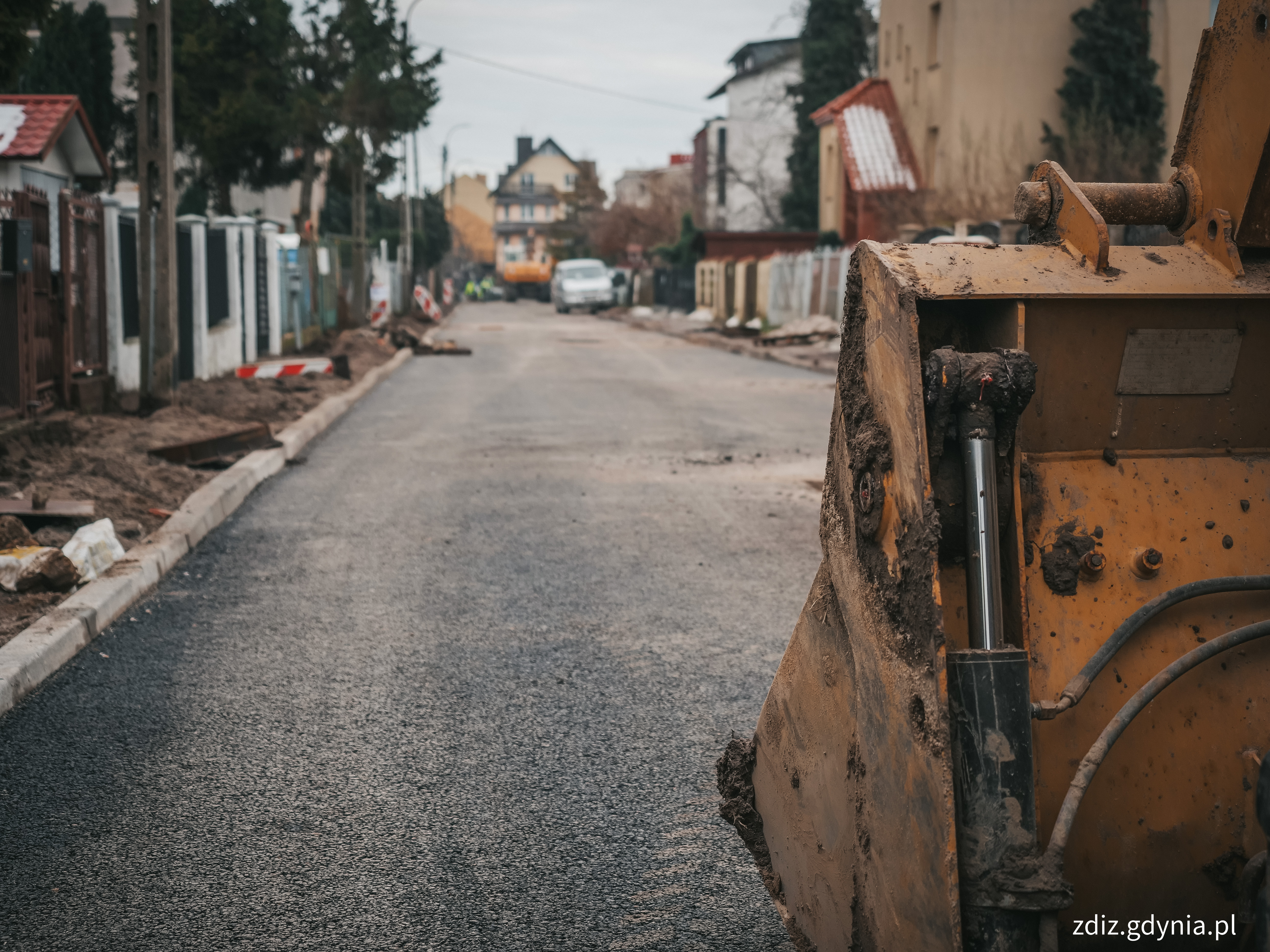 samoch&oacute;d na drodze, widoczna nowa nawierzchnia, w tle budynki