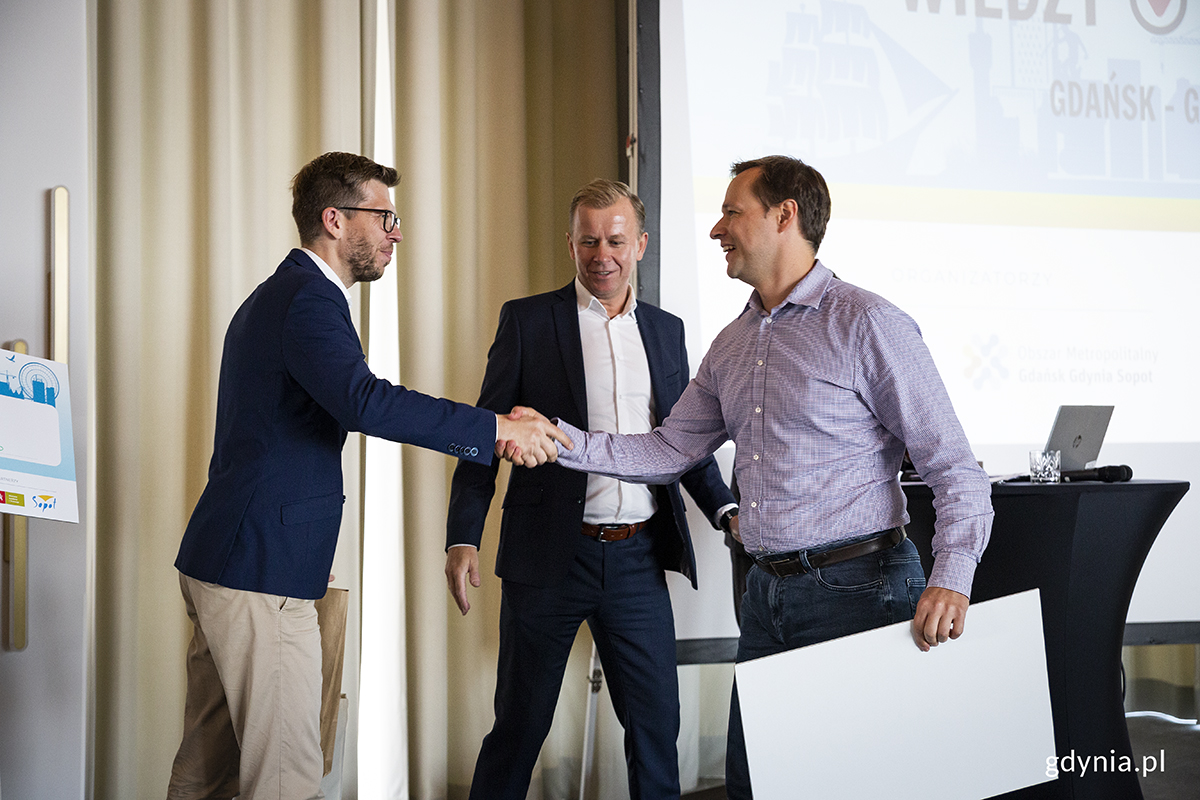 Trzej mężczyźni na scenie, jeden z nich odbiera nagrodę i gratulacje.