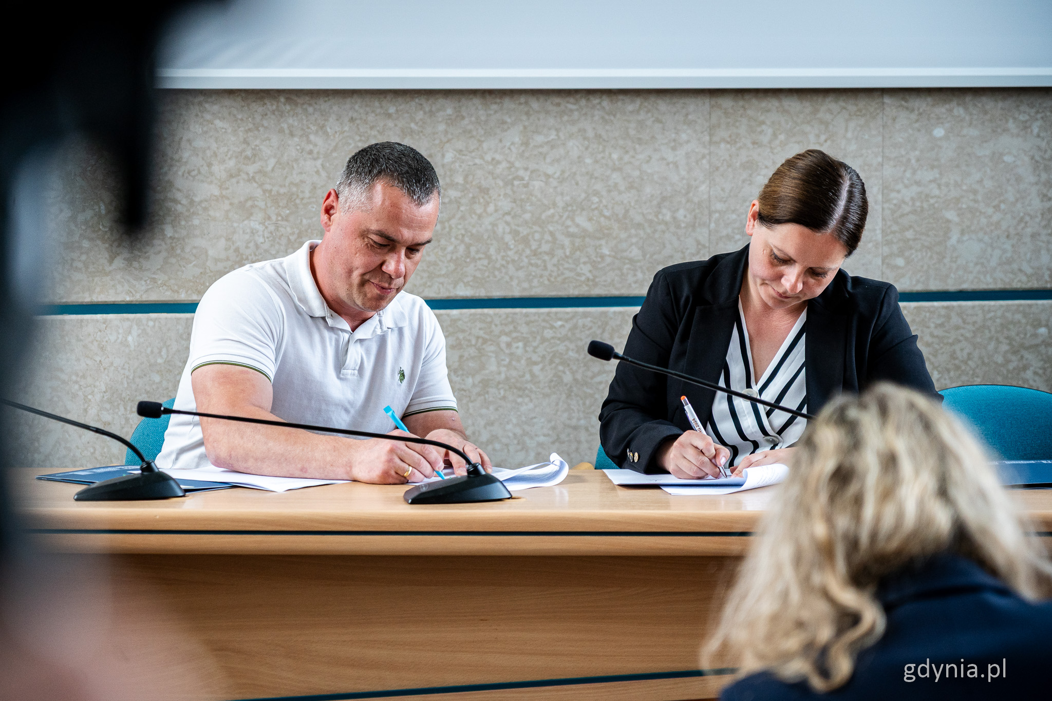 Sala obrad Rady Miasta Gdyni, przy stole siedzi mężczyzna w białej koszulce i Aleksandra Kosiorek, podpisują umowy