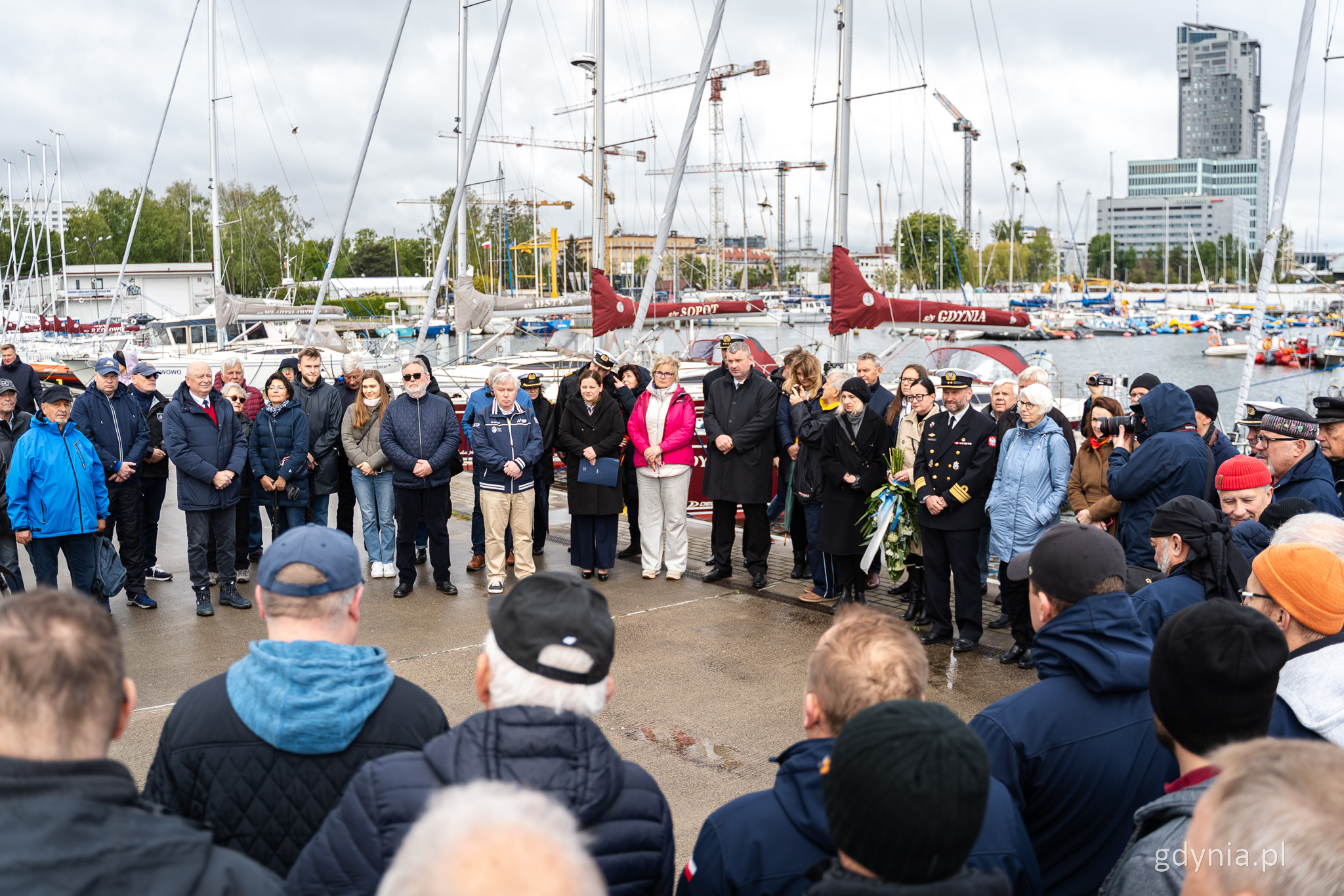 Nabrzeże w marinie Gdynia, grupa ludzi stoi w okręgu, uczestniczą w uroczystości, w tle marina, jachty, falochron