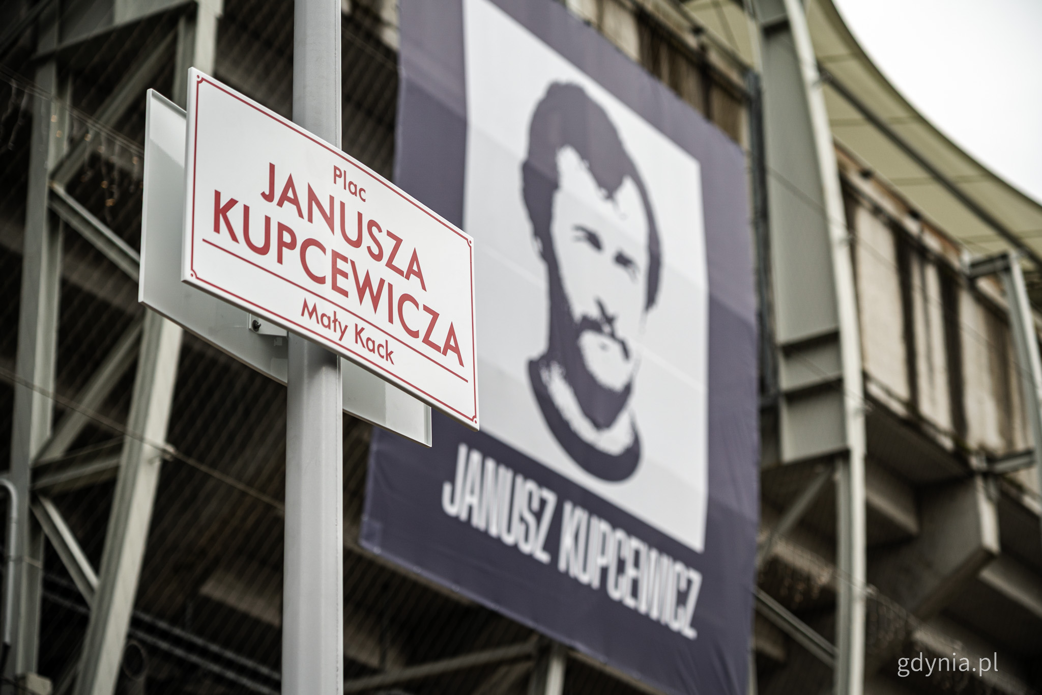 Tablica z napisem "plac Janusza Kupcewicza Mały Kack", w tle baner ze zdjęciem Janusza Kupcewicza na betonowej trybunie