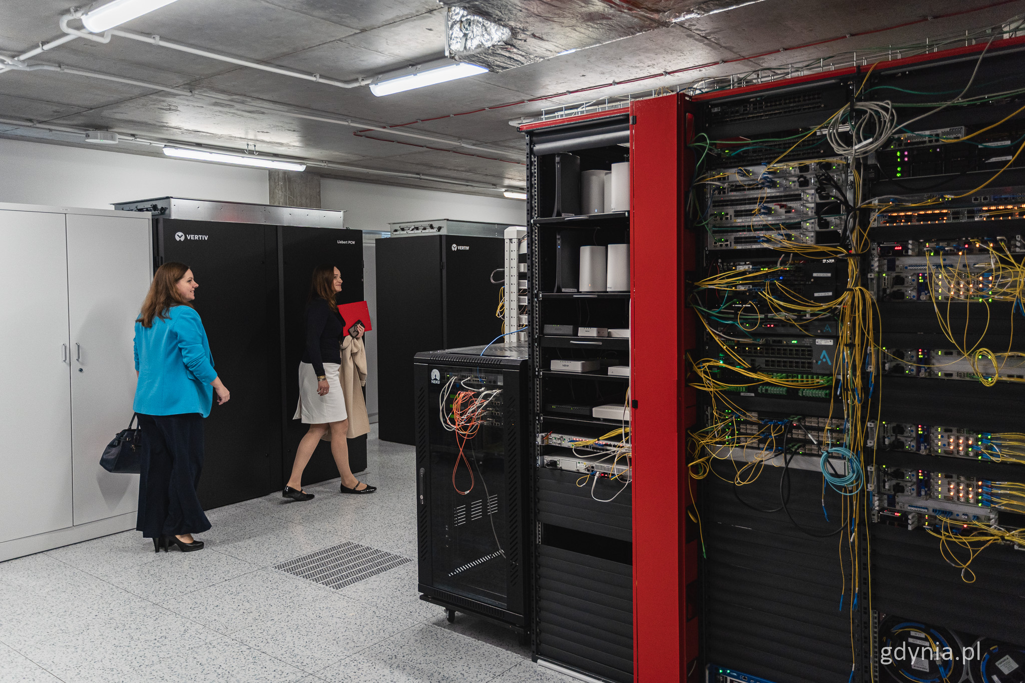 Pomieszczenie ze sprzętem, szafy ze sprzętem elektrycznym i kablami, z lewej obok urządzeń przechodzi Aleksandra Kosiorek i Izabela Disterheft