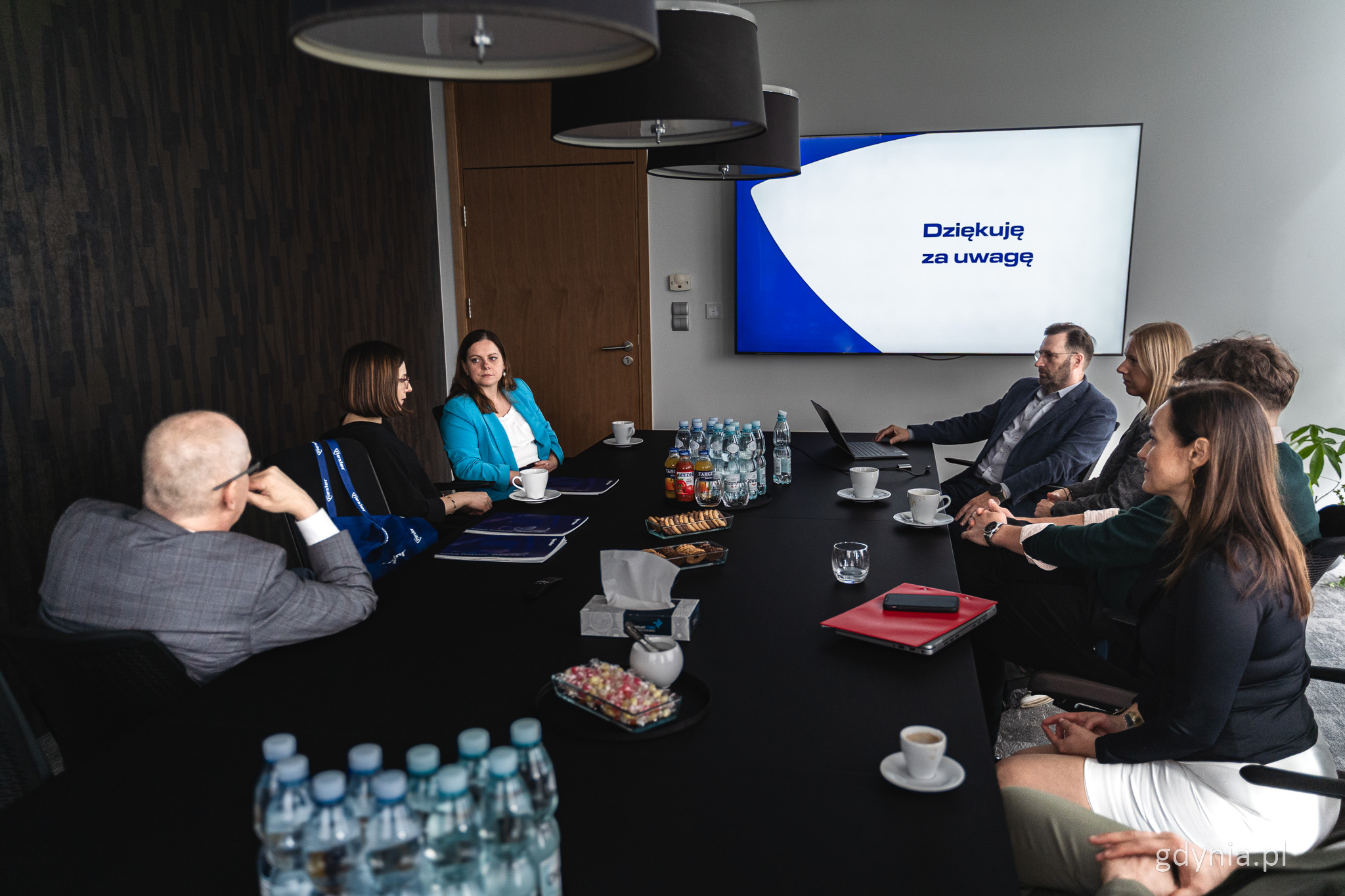 Sala konferencyjna, prezentacja wyświetlana na ekranie, kilka os&oacute;b siedzi dookoła stołu i rozmawia