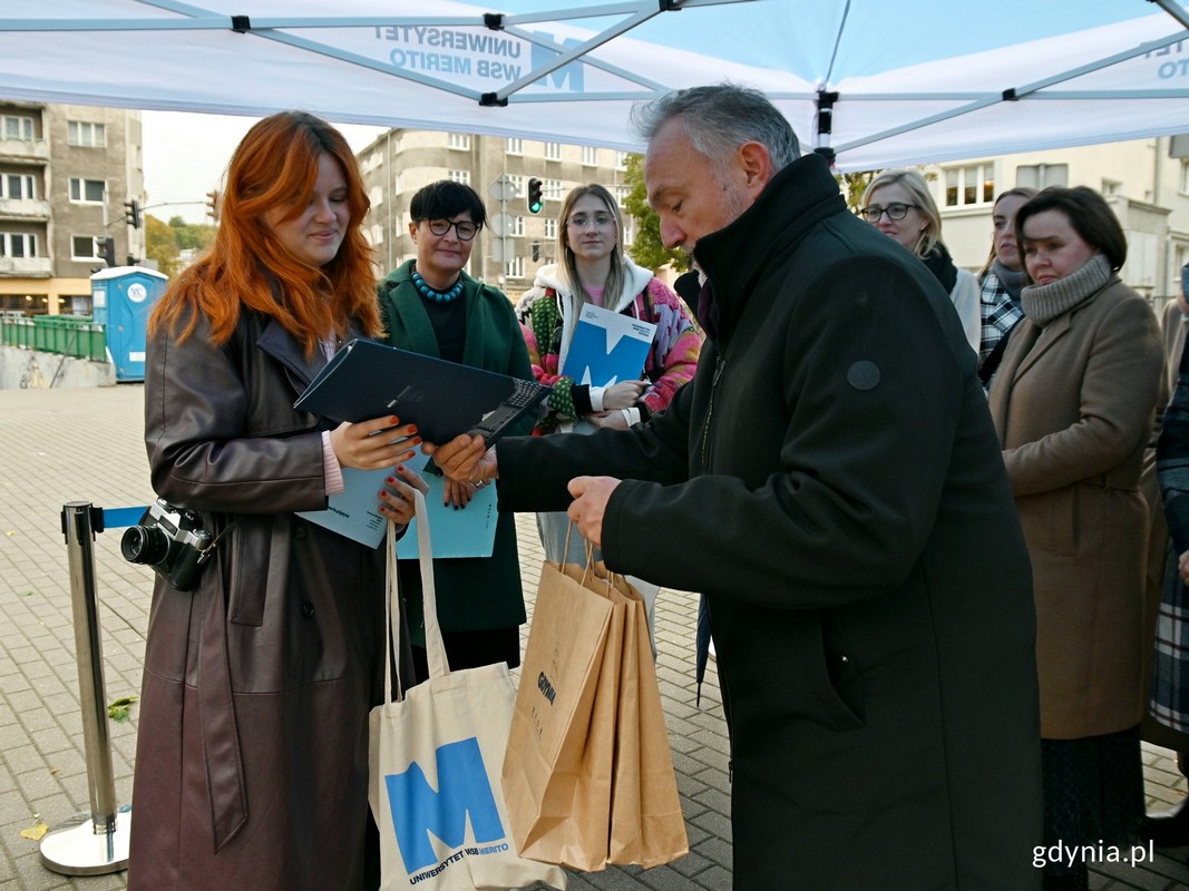 Prezydent Gdyni Wojciech Szczurek wręcza autorce zwycięskich projekt&oacute;w w konkursie "Uniwersytet WSB Merito maluje się dla Gdyni" - Kindze Kulpie - list gratulacyjny i torbę z drobnymi upominkami // fot. Magdalena Czernek