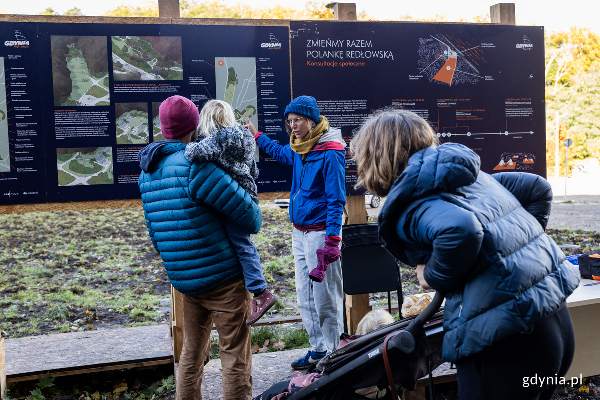 Kobieta przy tablicy informacyjnej na lesnej polanie opowiada rodzinie o planach zagospodarowania.
