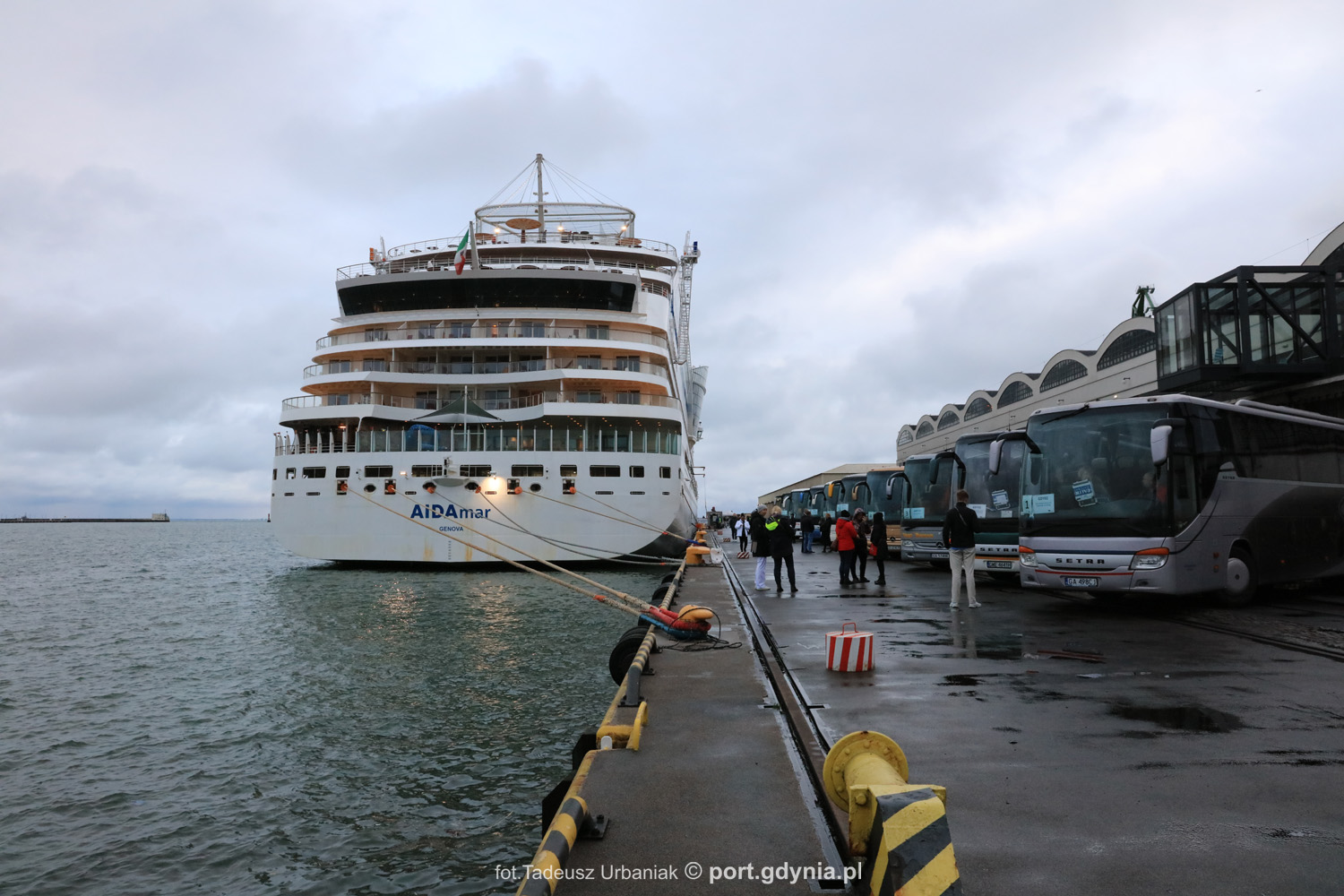Statek wycieczkowy zacumowany przy nabrzeżu, na kt&oacute;rym stoją ludzie i autokary.