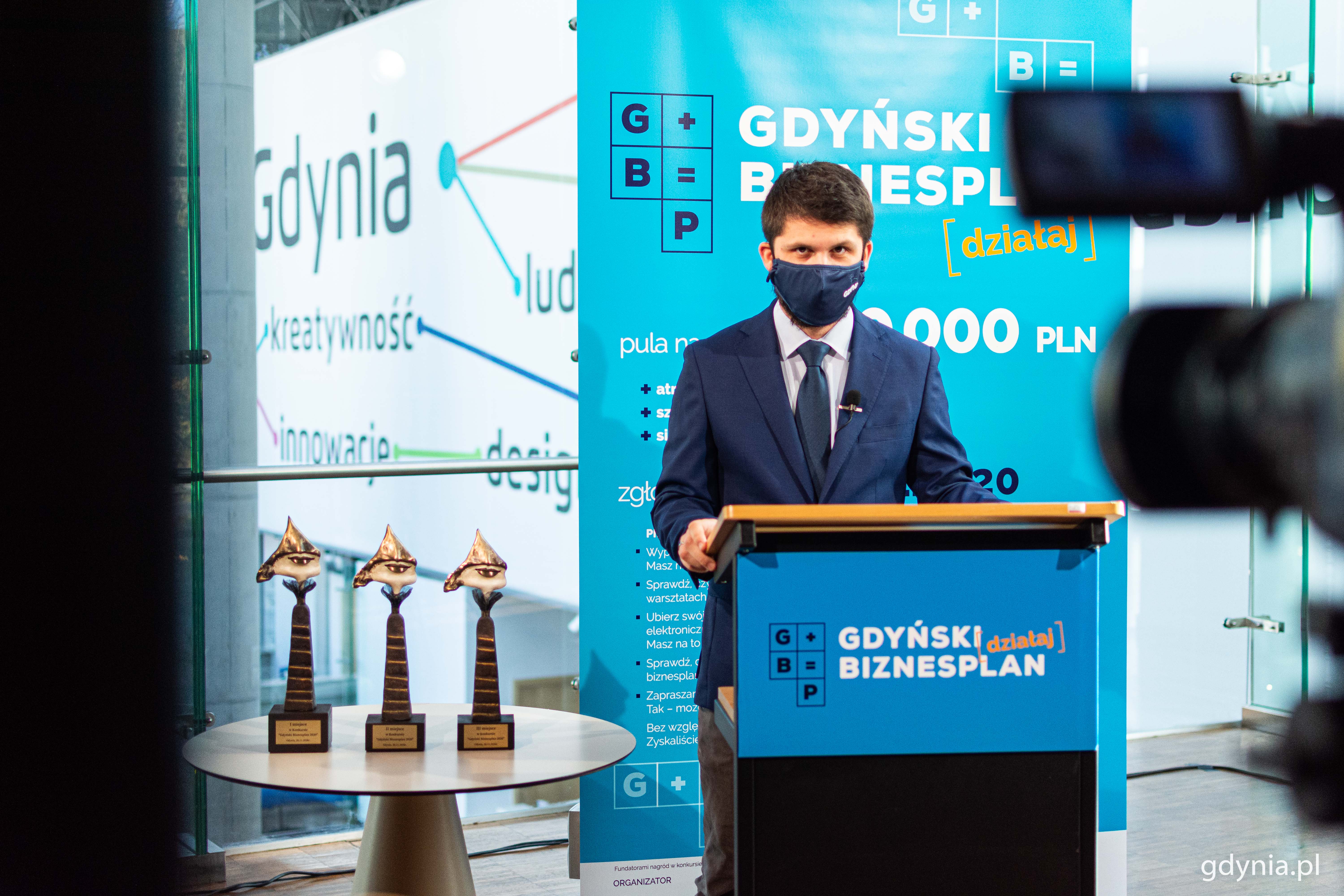 Zdjęcie z gali finałowej konkursu Gdyński Biznesplan, kt&oacute;ra w tym roku odbyła się online. Prowadzący stoi przed kamerą, za nim niebieska plansza z logiem konkursu. Po lewej stronie, na stoliku stoją statuetki dla laureat&oacute;w // fot. Kamil Złoch