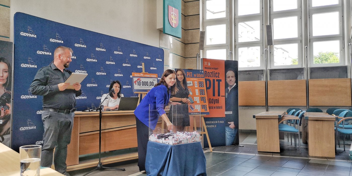 Sala obrad Rady Miasta w Urzędzie Miasta Gdyni, st&oacute;ł, szklana kula z losami, prowadzący i losująca w kuli prezydent Gdyni Aleksandra Kosiorek, ścianki reklamowe