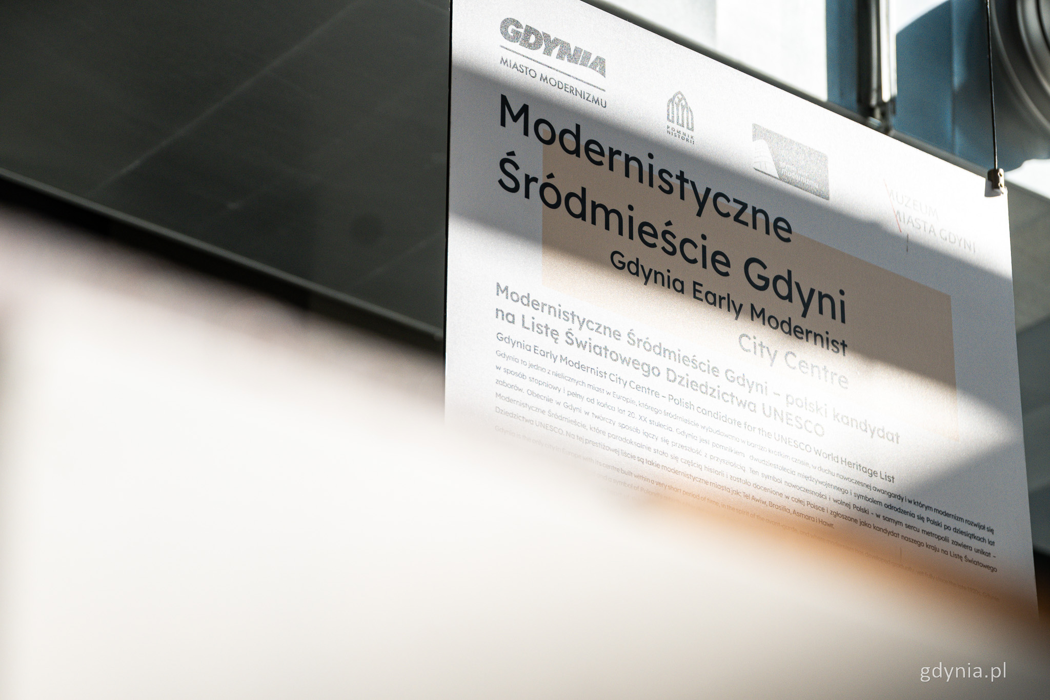 Wystawa, wisząca tablica dotycząca gdyńskiego modernizmu, zdjęcia, napisy, w tle ściany i przeszklenia