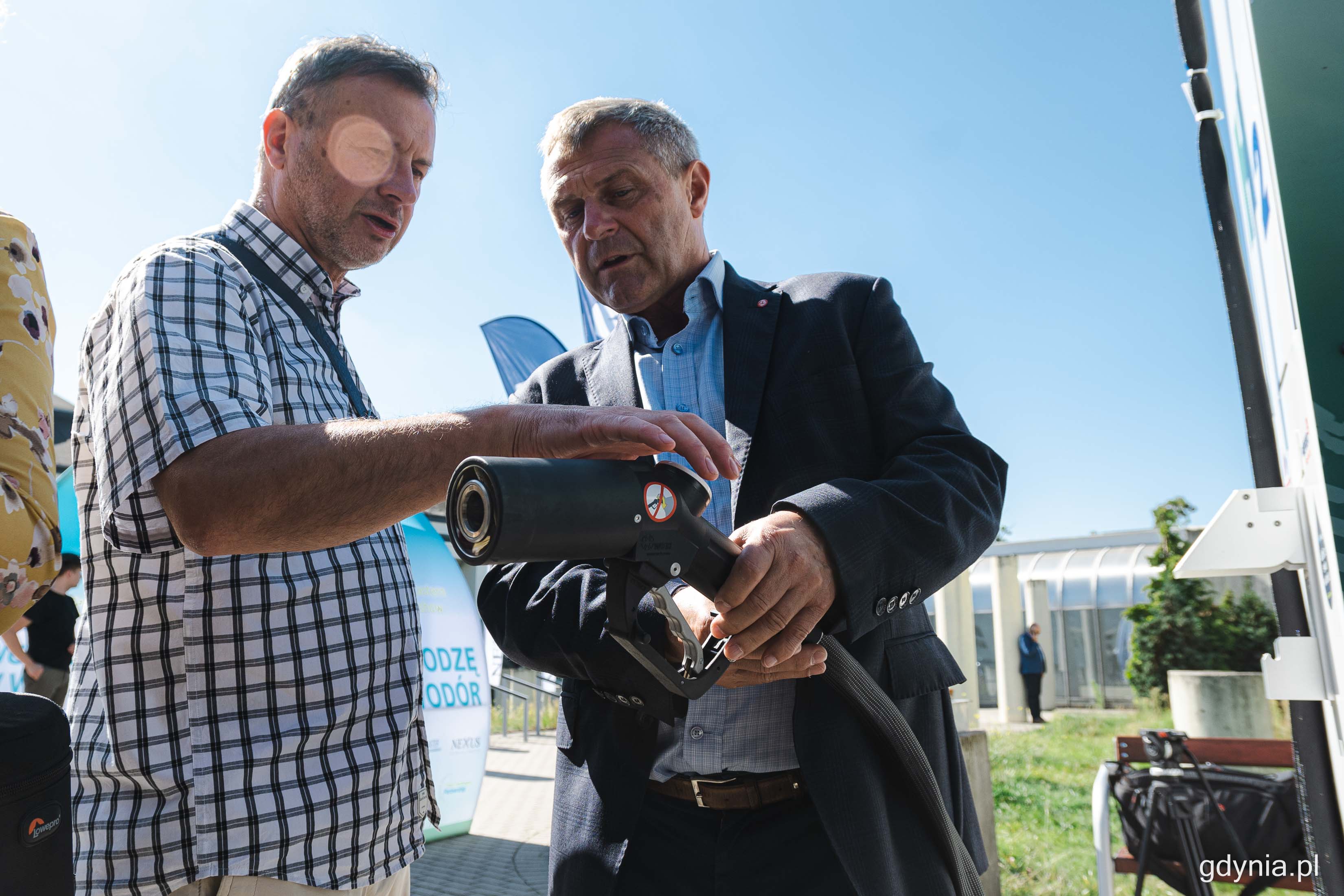 Dw&oacute;ch mężczyzn, jeden trzyma pistolet do tankowania wodoru, drugi przygląda się urządzeniu, w tle budynki PPNT Gdynia, słoneczny dzień