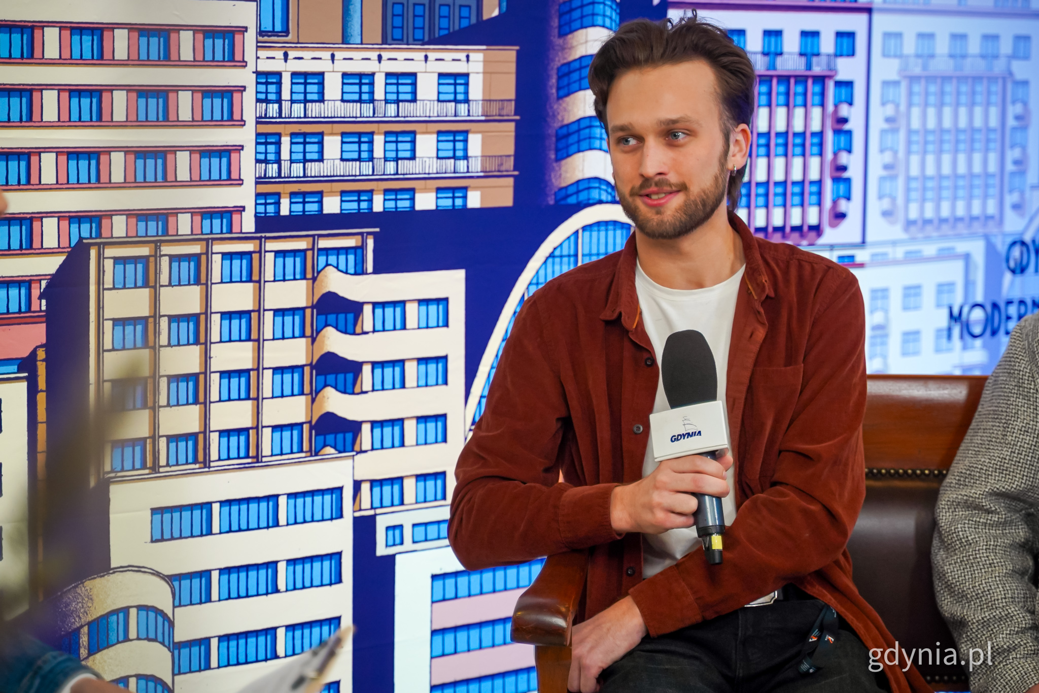 Piotr Bańkowski siedzi na ławie, trzyma mikrofon