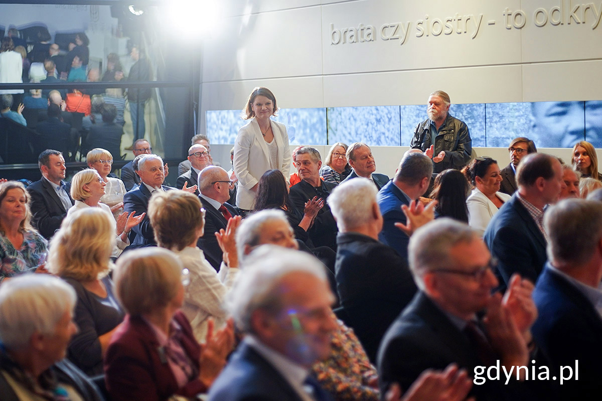 Tłum ludzi na sali, jedna kobieta stoi.