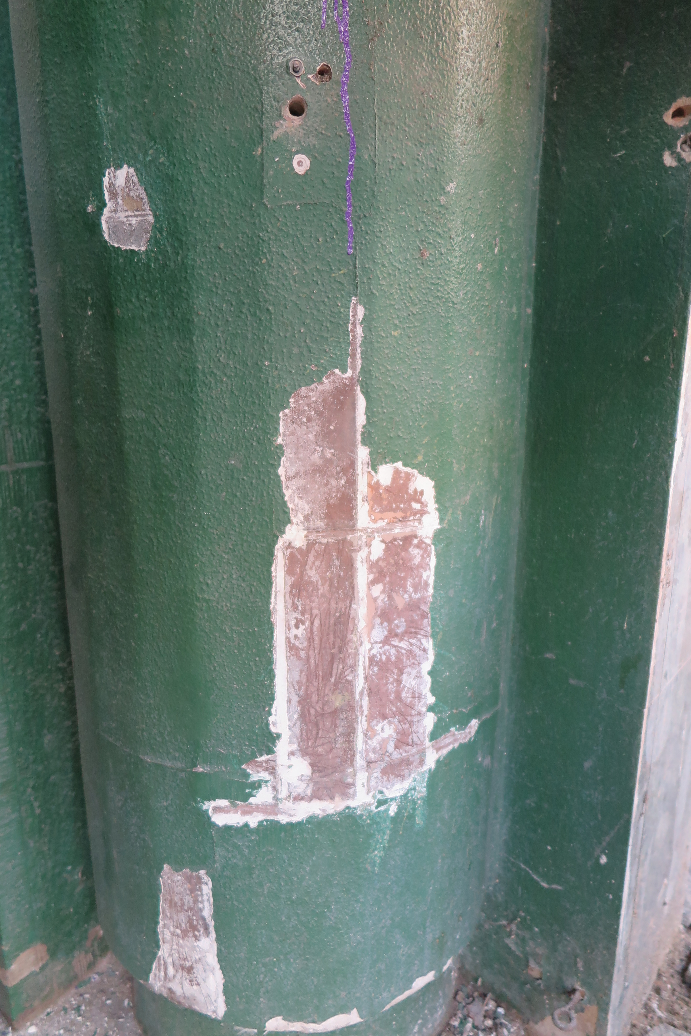 Szczeg&oacute;ł filaru flankującego wejście do budynku &ndash; spod warstwy zielonej farby wyłania się oryginalne wykończenie brązowymi kształtkami ceramicznymi.