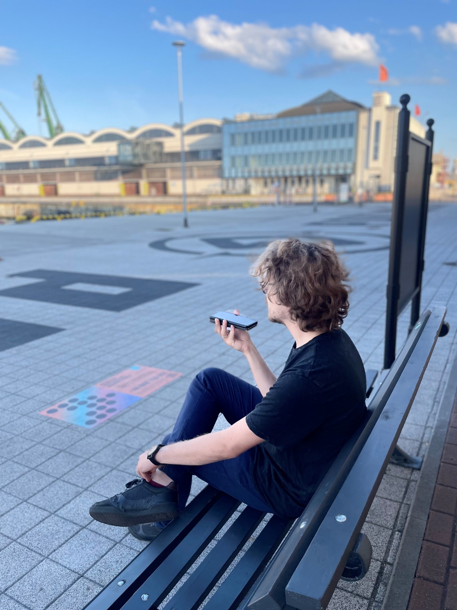 Mężczyzna z telefonem kom&oacute;rkowym w dłoni siedzi na ławce. W tle widać budynek Muzeum Emigracji