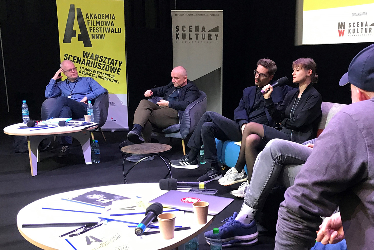 Kilka os&oacute;b siedzi na fotelach przy stolikach podczas debaty.