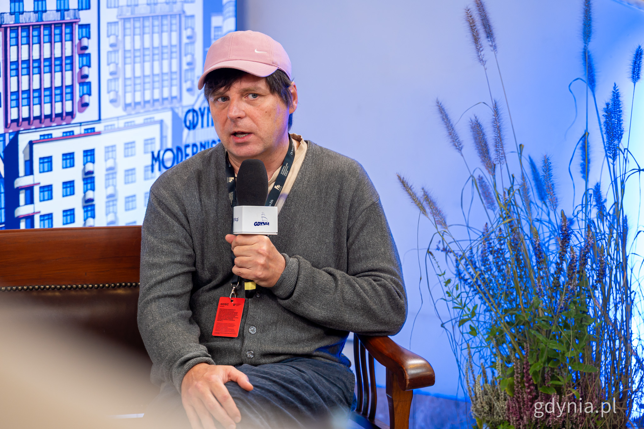 Mężczyzna w r&oacute;żowej czapce i szarym swetrze siedzi na kanapie, m&oacute;wi do mikrofonu. Sebastian Pańczyk