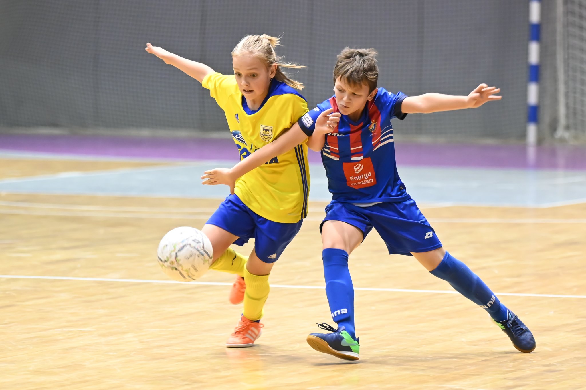 Na zdjęciu dw&oacute;ch młodych chłopc&oacute;w - jeden w ż&oacute;łtej, drugi w granatowej koszulce - walczących o piłkę w hali. 