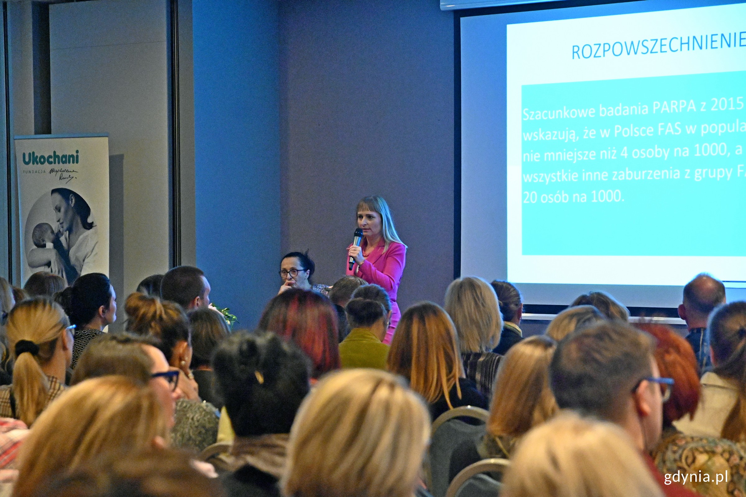 kobieta przemawiająca do dużej grupy zebranych ludzi w miejscu konferencji
