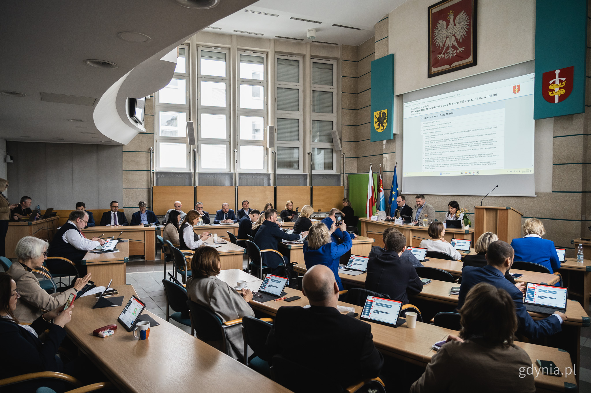 Sala obrad Rady Miasta w Urzędzie Miasta Gdyni, radni siedzą w ławach, na ekranie wyświetlany tekst, na środku prezydium rady