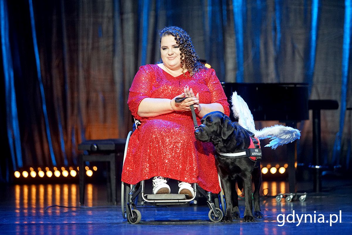 Kobiet w czerwonej sukni siedząca na w&oacute;zku inwalidzkim, obok niej czarny pies przewodnik 