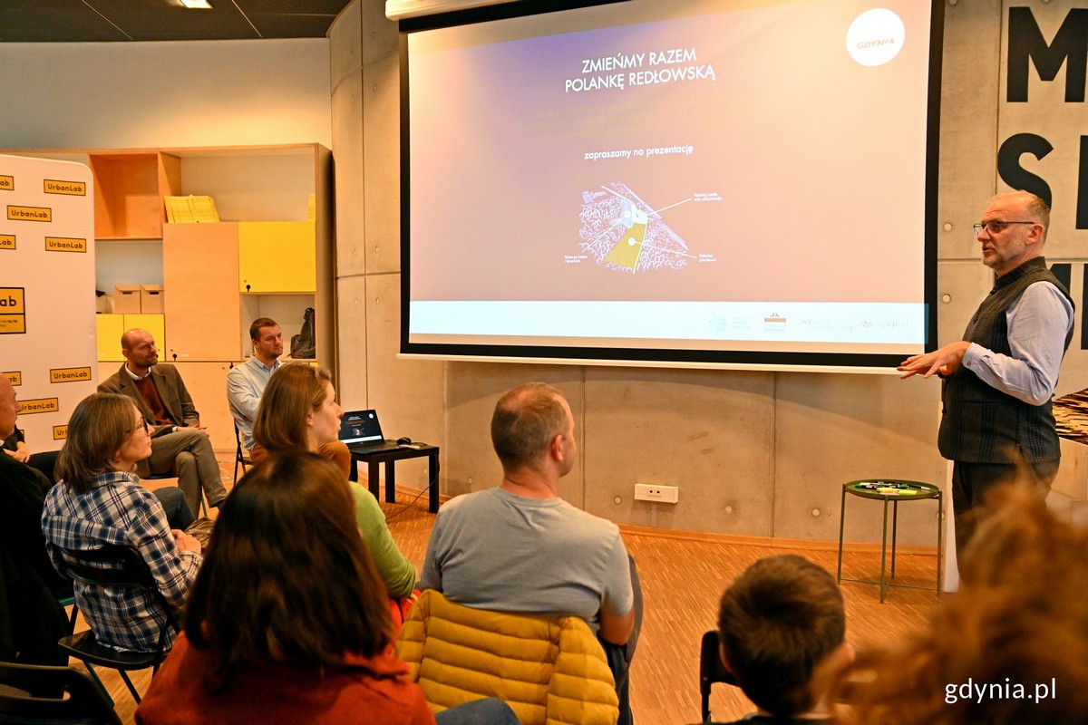 Mężczyzna stoi i przy ekranie i opowiada, na sali siedzą ludzie.