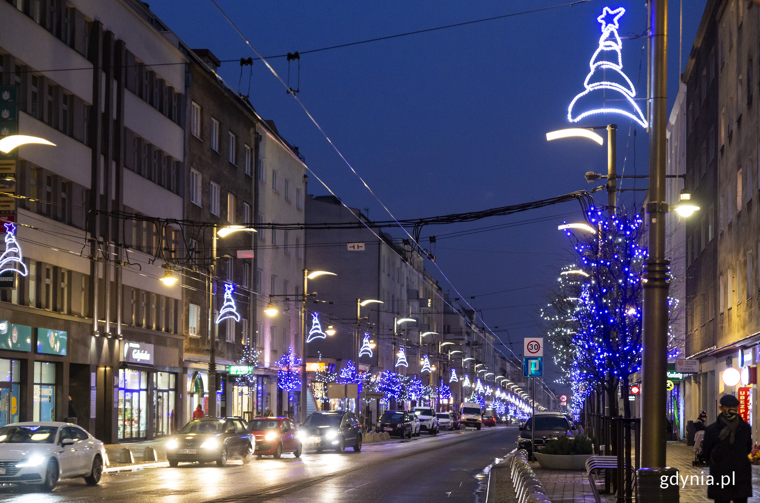 Ulica Świętojańska. Latarnie przyozdobione są świątecznymi choinkami świecącymi na niebiesko. Drzewa obok latarni ozdobione są łańcuchami świetlnymi, kt&oacute;re r&oacute;wnież świecą na niebiesko. // fot. Przemysław Kozłowski