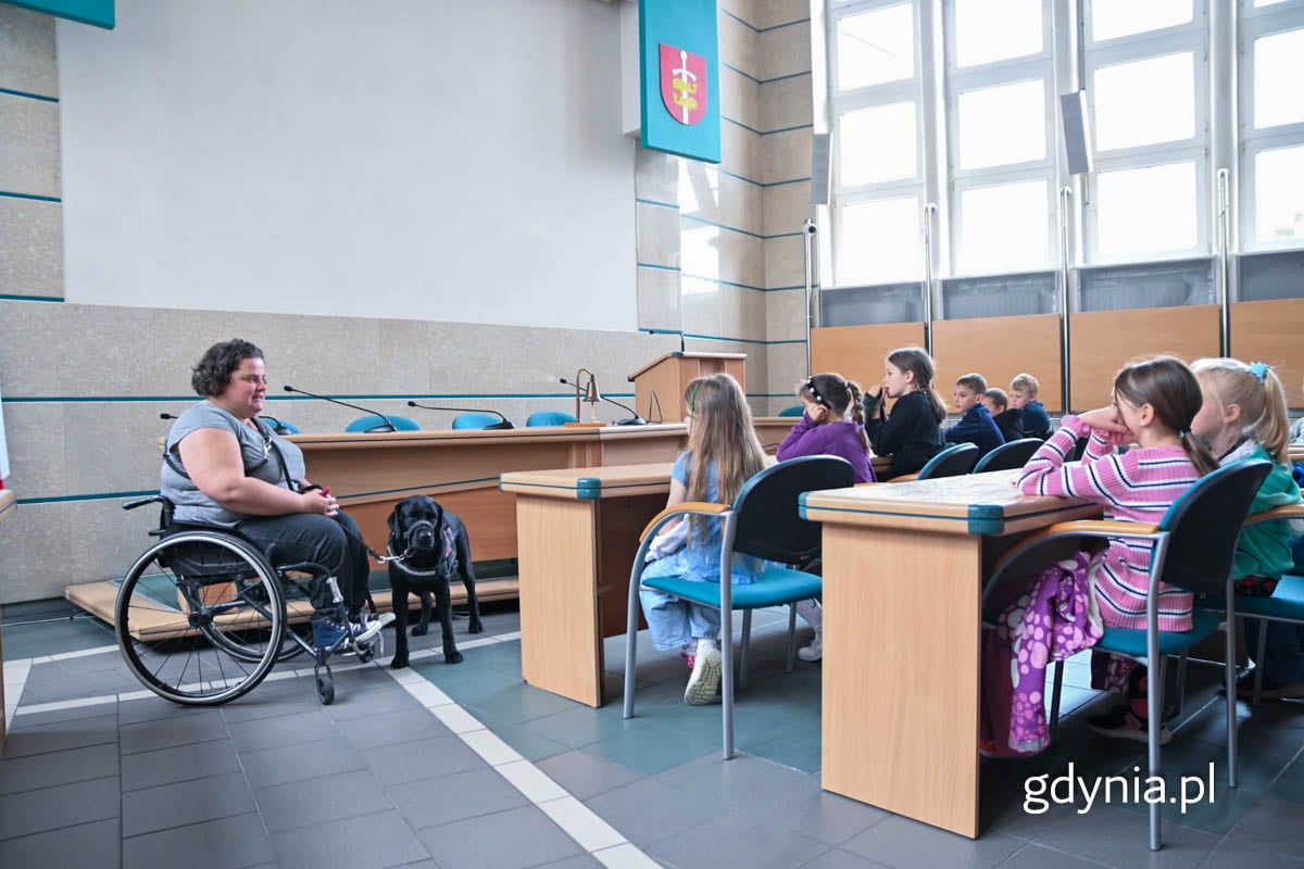 Kobieta siedząca na w&oacute;zku inwalidzkim, obok niej labrador - pies przewodnik oraz dzieci siedziące przy stołach na sali obrad Rady Miasta Gdyni.