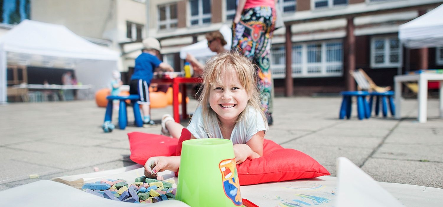 Uśmiechnięta dziewczynka podczas zabawy na betonowej posadzce. W tle kobieta z dzieckiem i instruktorem, znajdujący się przy stole.