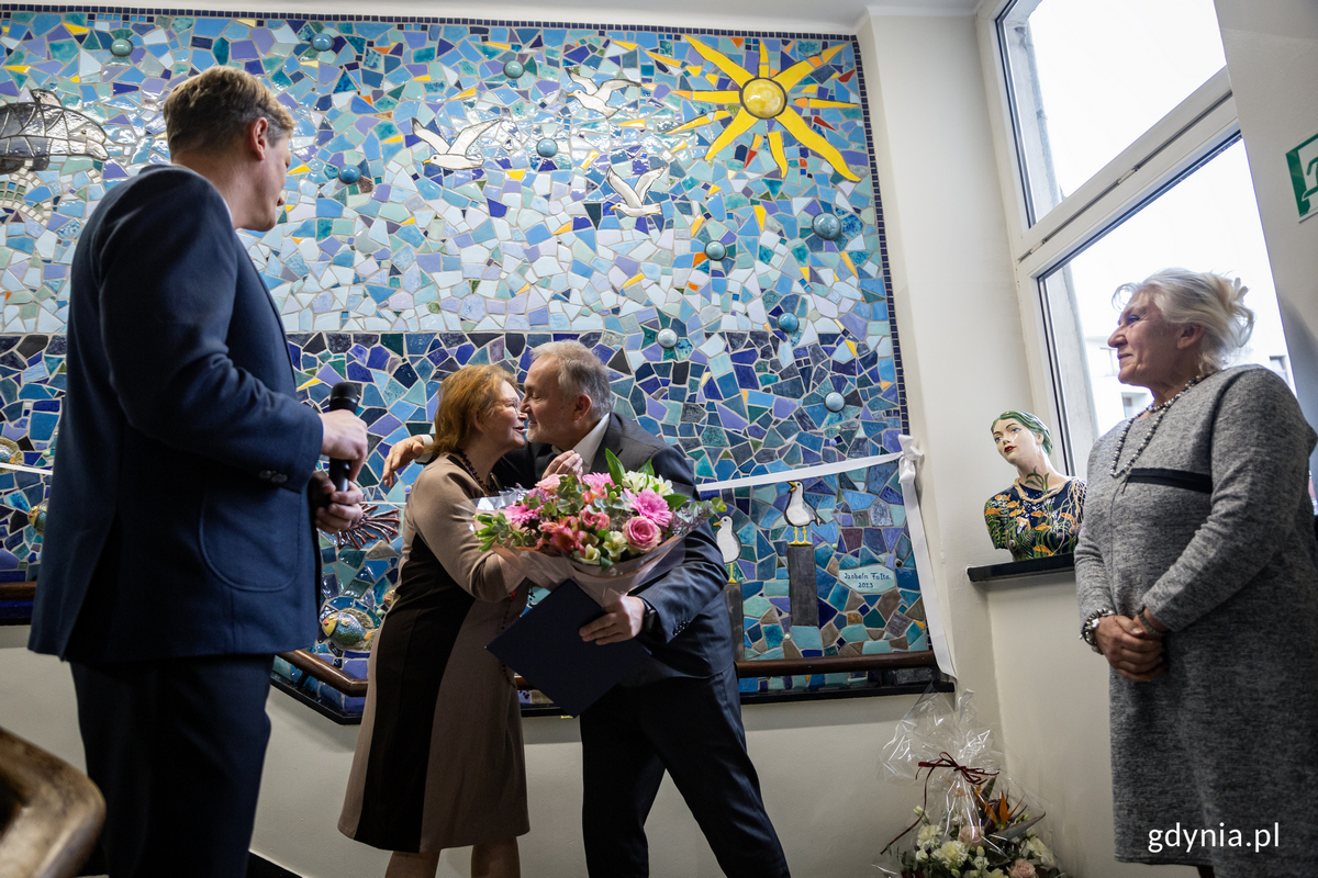 Mężczyzna wręcza kwiaty kobiecie, w tle mozaika.