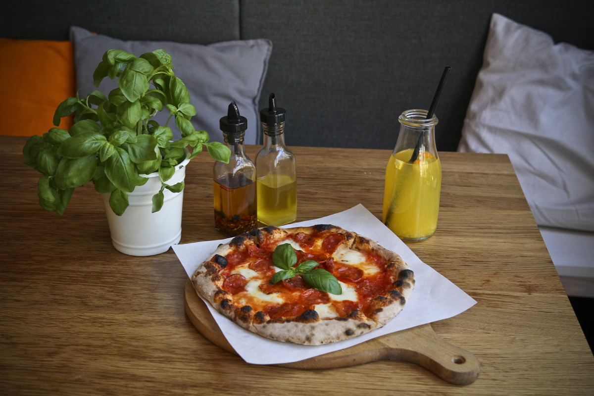 Na środku stołu, na drewnianej desce leży pizza. Obok stoi doniczka z bazylią, dwie szklane budelki z oliwą z oliwek oraz szklana butelka z lemoniadą. W tle widoczne są siedzenia i poduszki. // fot. Przemysław Kozłowski
