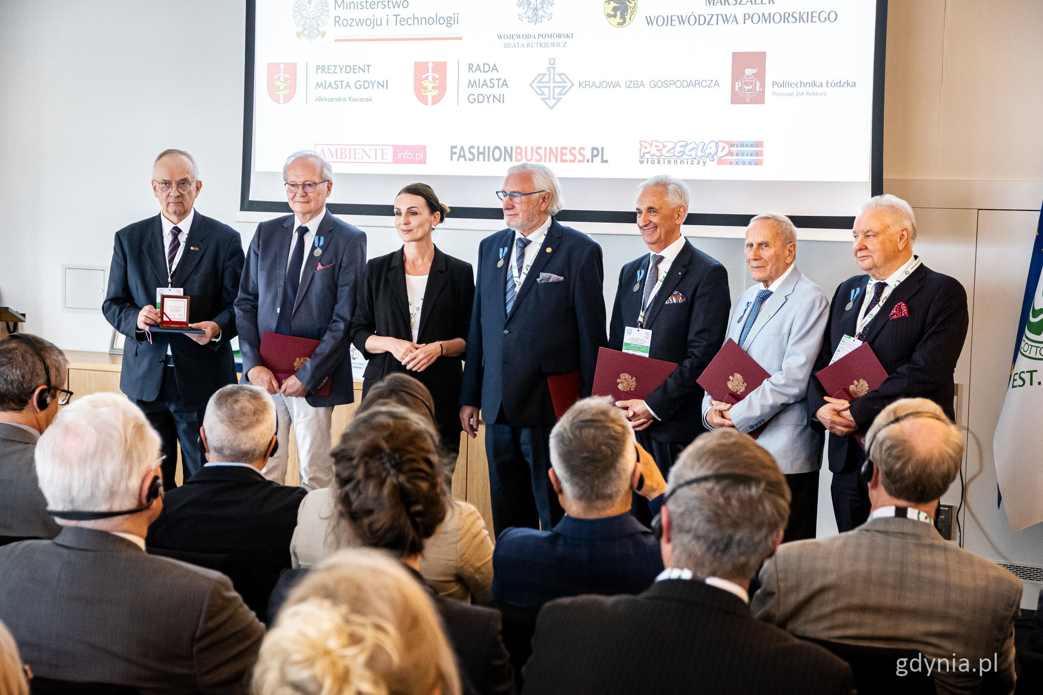 Sala konferencyjna, ekran, grupa os&oacute;b w formalnych strojach pozuje do zdjęcia, mają przypięte ordery