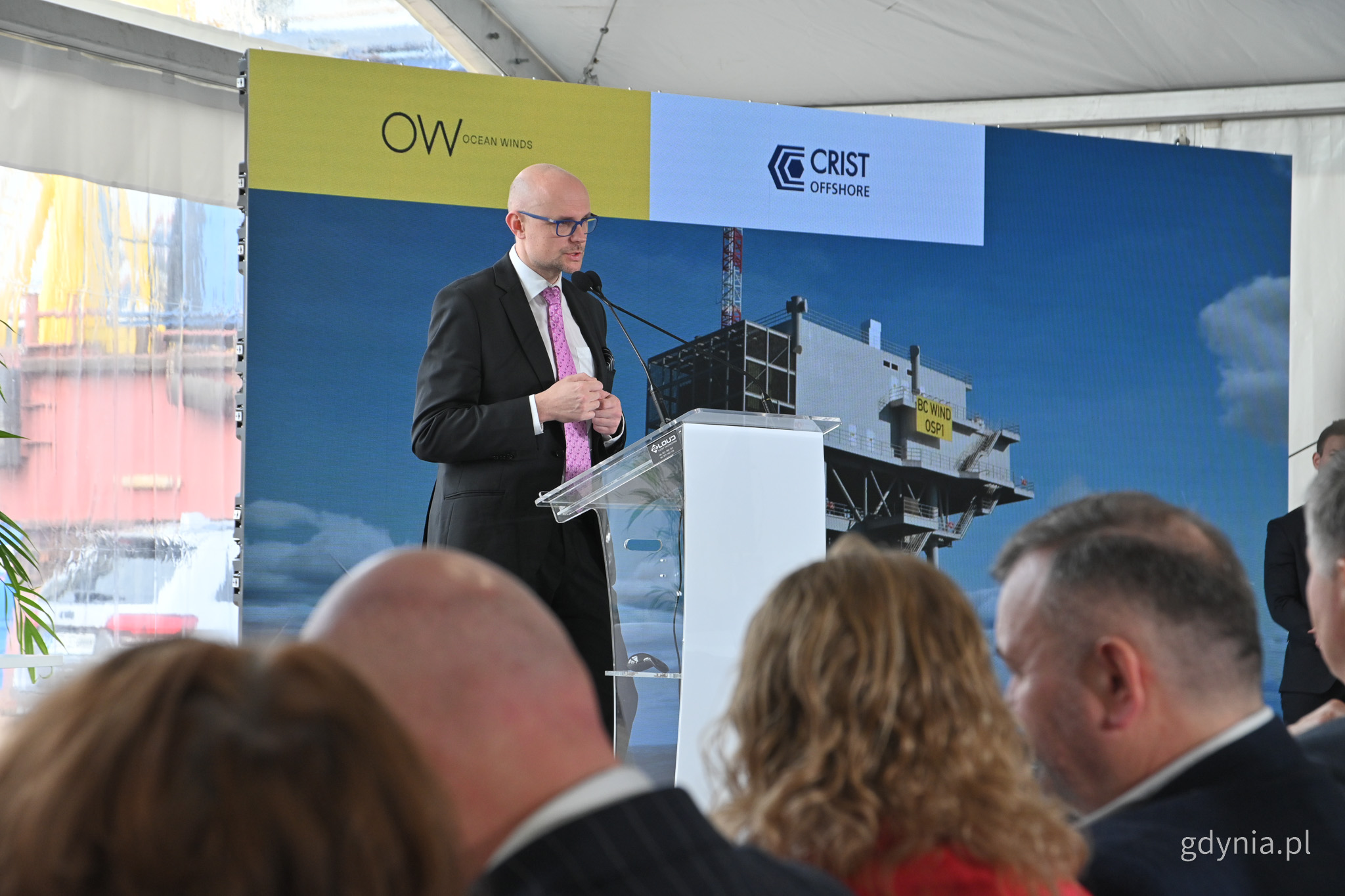 Scena w namiocie konferencyjnym, przy m&oacute;wnicy stoi Bartłomiej Austen, w tle ekran z logotypami firmy