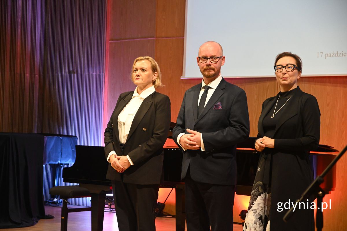 Dwie kobiety i mężczyzna, ubrani elegancko, stoją na scenie