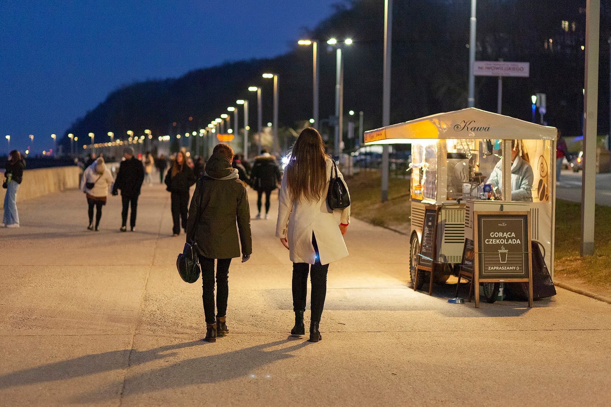 Wiecz&oacute;r, bulwar w Gdyni, grupa os&oacute;b spaceruje, na poboczu stoi rowerowa kawiarnia