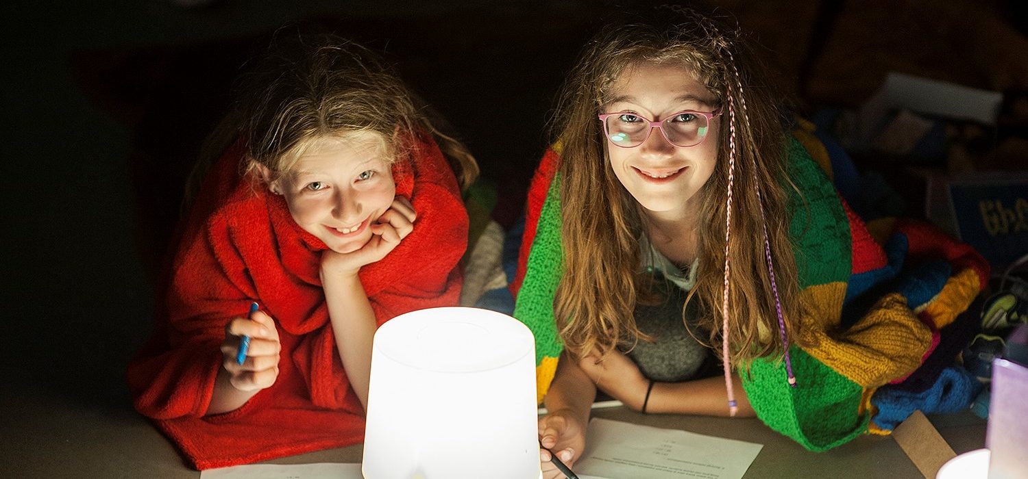 Dw&oacute;jka dzieci pod kocami, trzymająca w rękach długopisy i kredki. Oświetla ich lampa. 