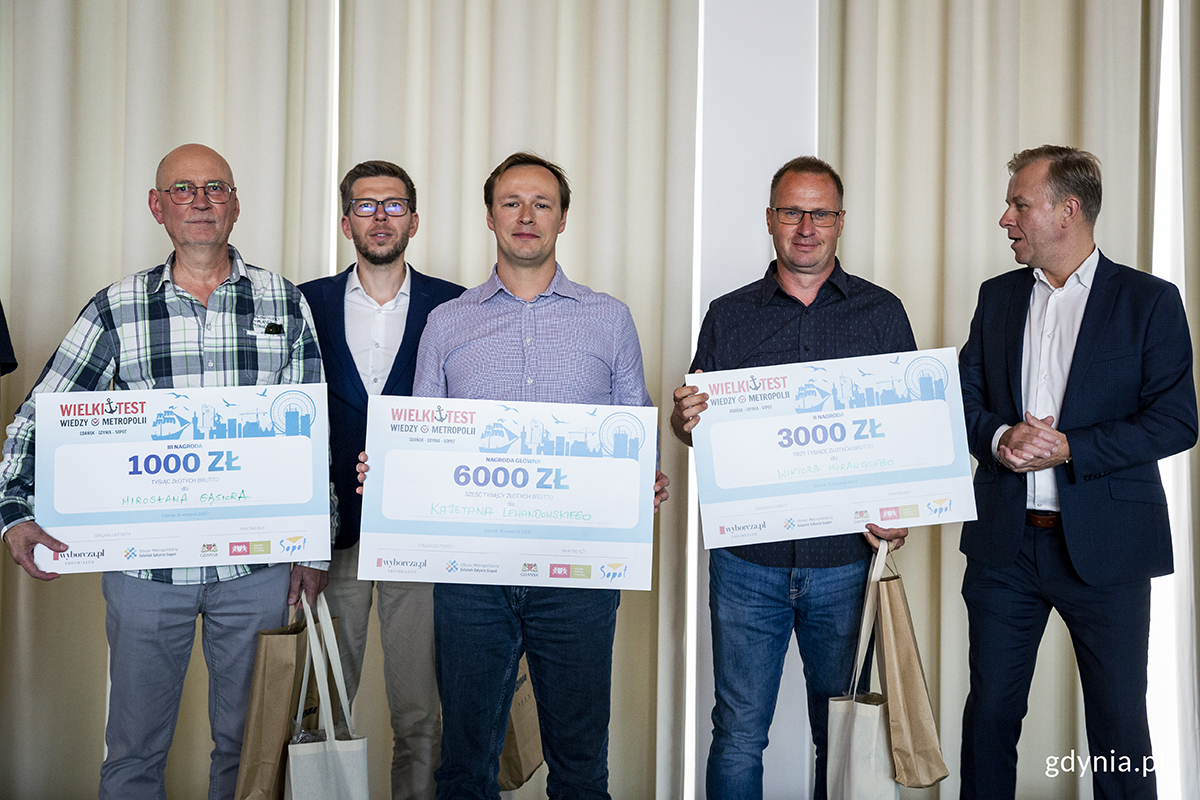 Pięciu mężczyzn na scenie, trzech z nich trzyma tablice z kwotami wygranych pieniędzy.