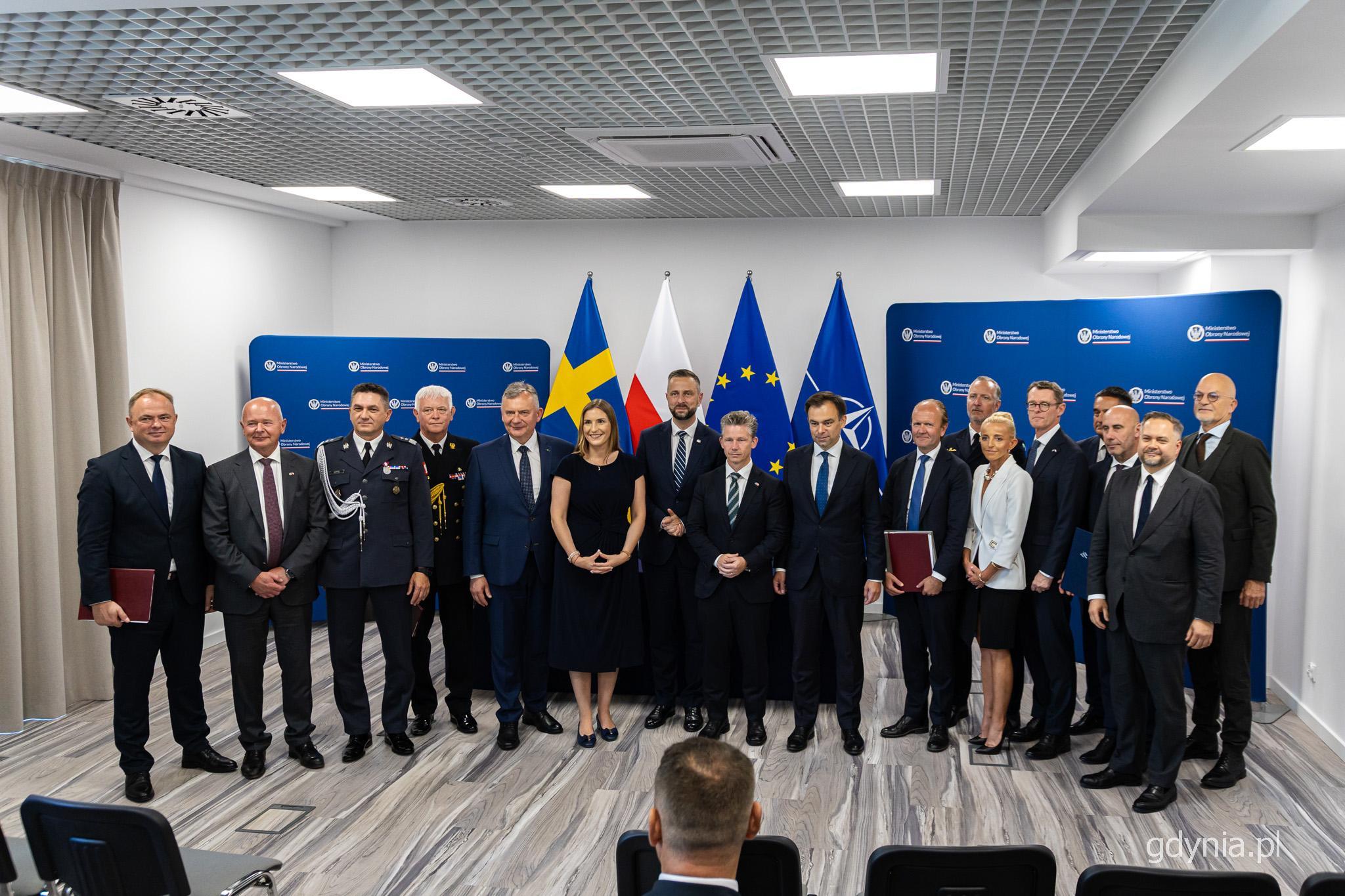 Sala konferencyjna, grupa przedstawicieli rządu Polski i Szwecji pozuje do zdjęcia, w tle ścianki z logotypami ministerstw