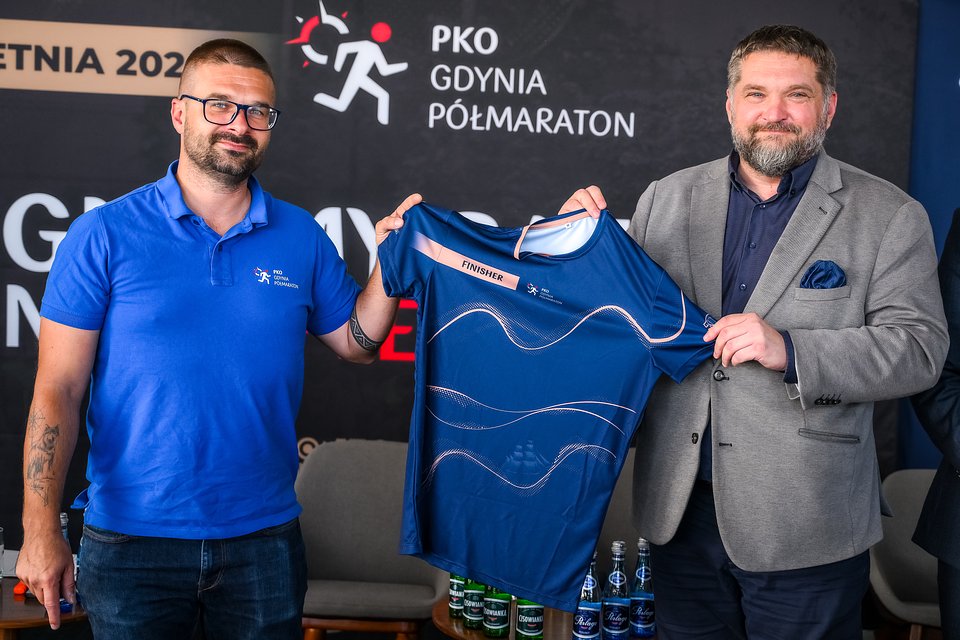 Michał Drelich (z lewej) i Tadeusz Szemiot (z prawej) - przewodniczący Rada Miasta Gdyni, prezentują oficjalną koszulkę finiszer'a PKO Gdynia P&oacute;łmaratonu 2026